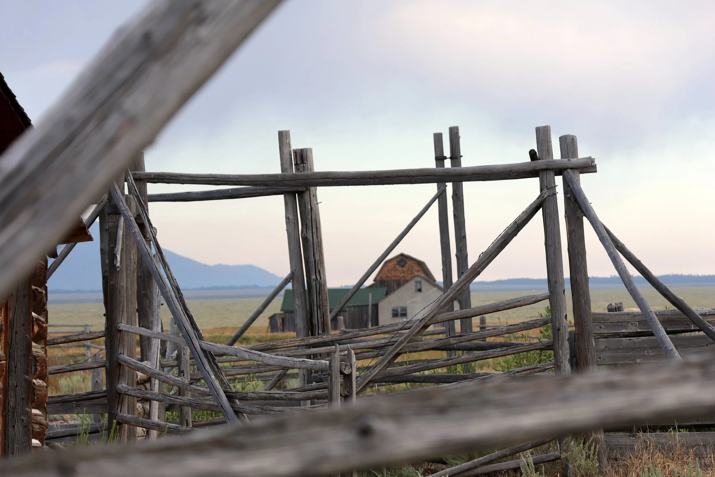 View of a wooden fence with a rural house, fields, and mountains in the background under a cloudy sky, seen through a weathered wooden structure.
