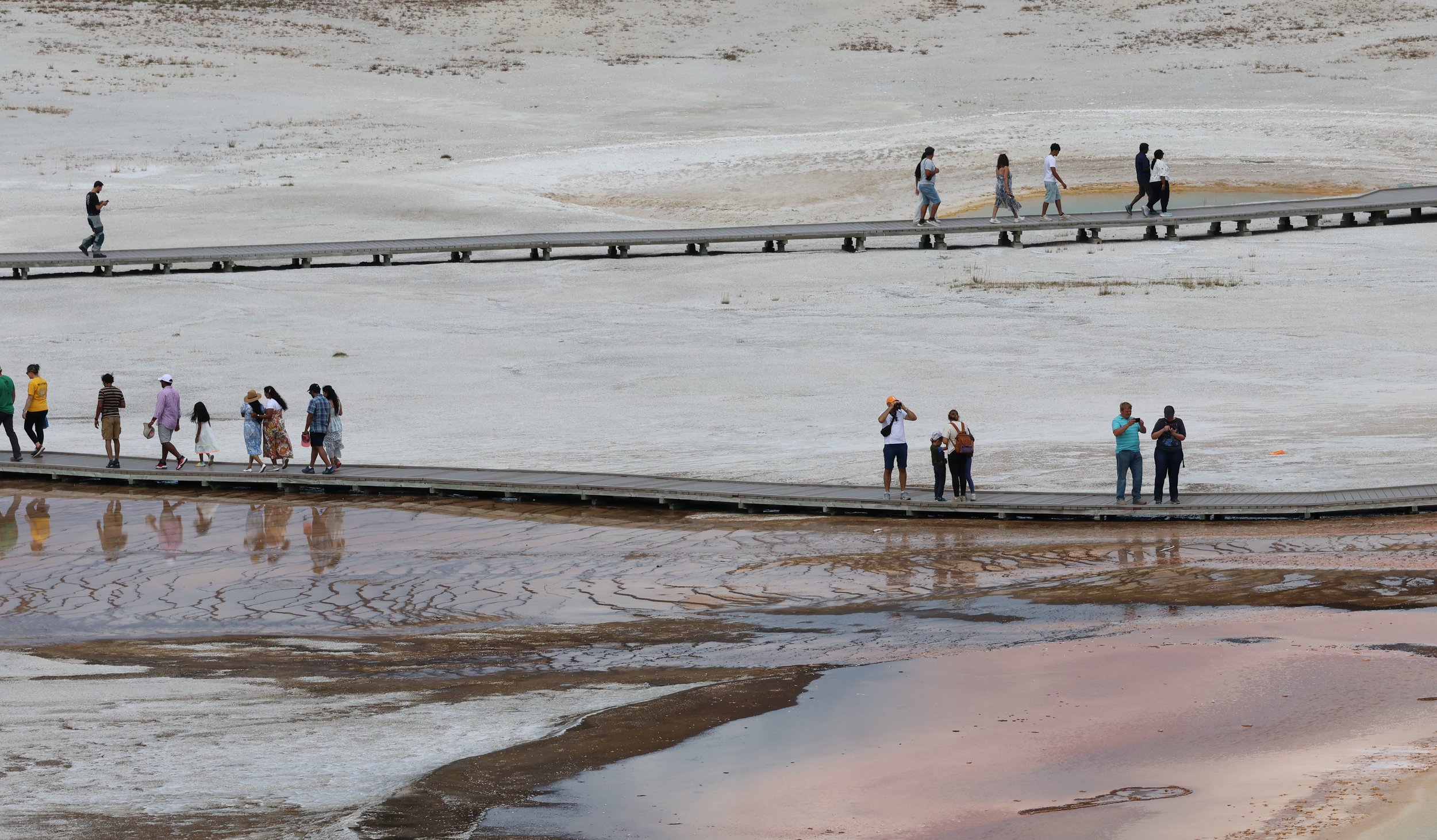 People walking on wooden pathways across a white salt flat with cracked salt crust and salt deposits in the foreground.