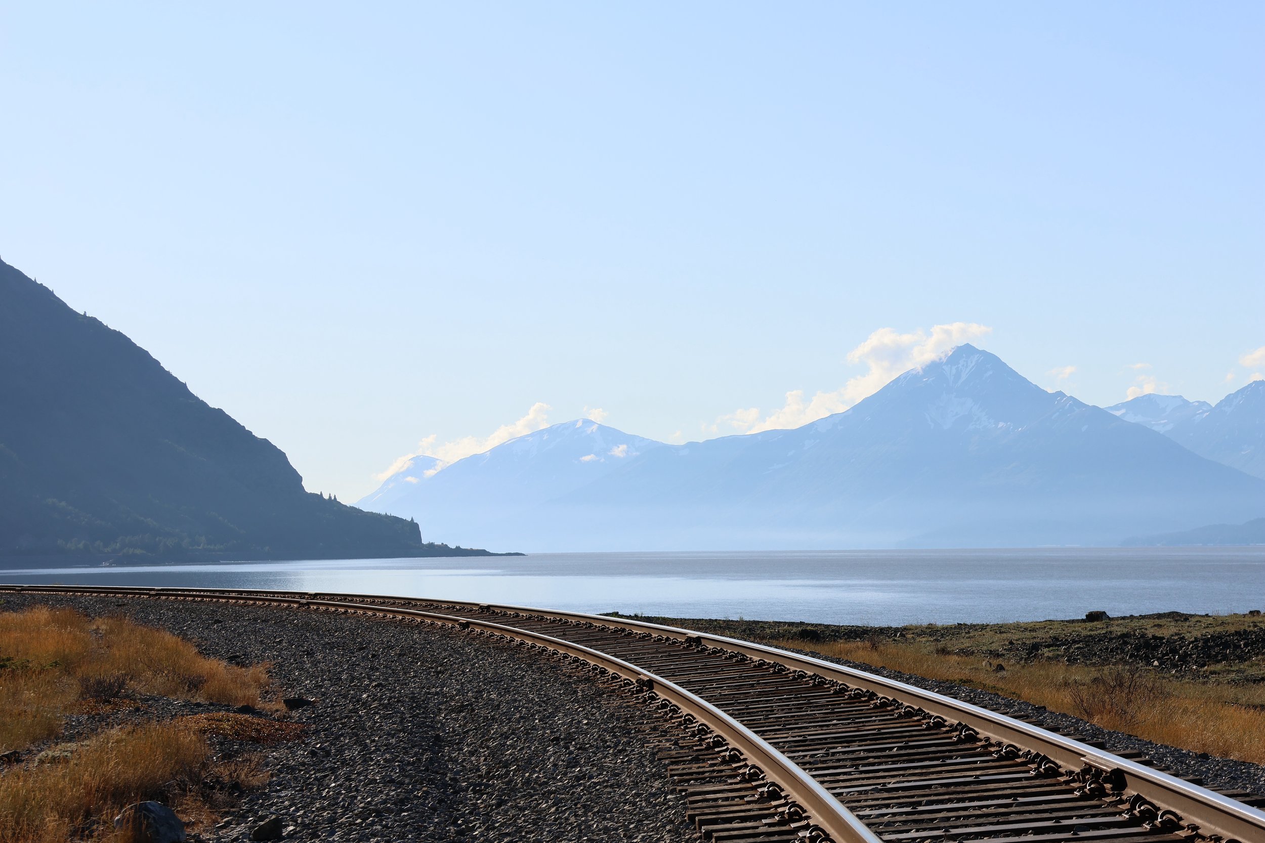 A landscape featuring snowy mountain peaks in the background, a large body of water, and railroad tracks in the foreground along the shoreline.