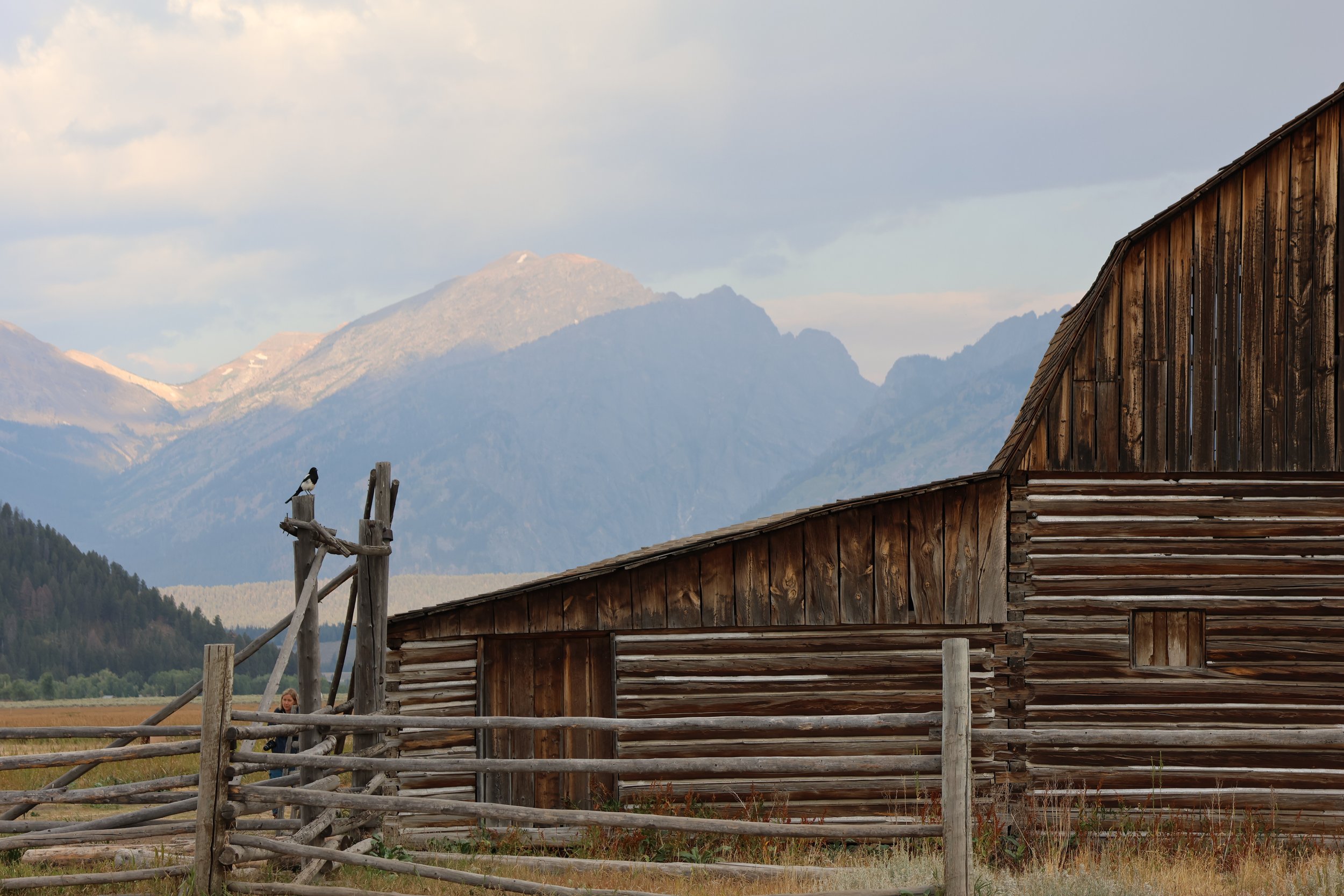 A rustic wooden barn with a mountainous landscape in the background, clouds in the sky, and a person visible near a wooden fence.