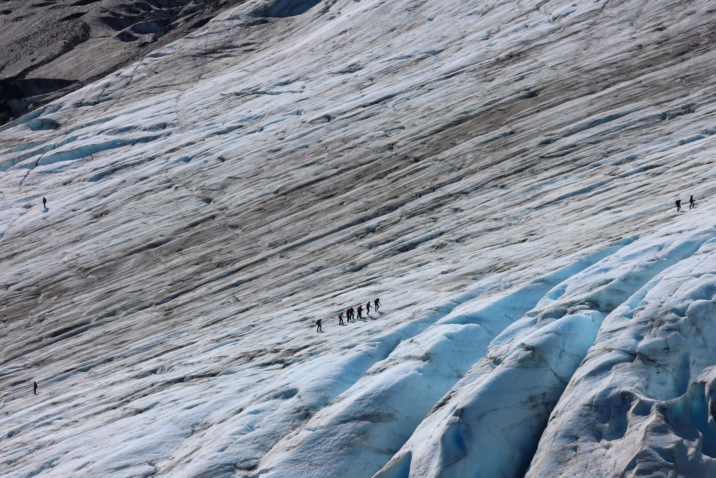 A glacier with visible ice crevasses and cracks, with a group of hikers walking on the surface.