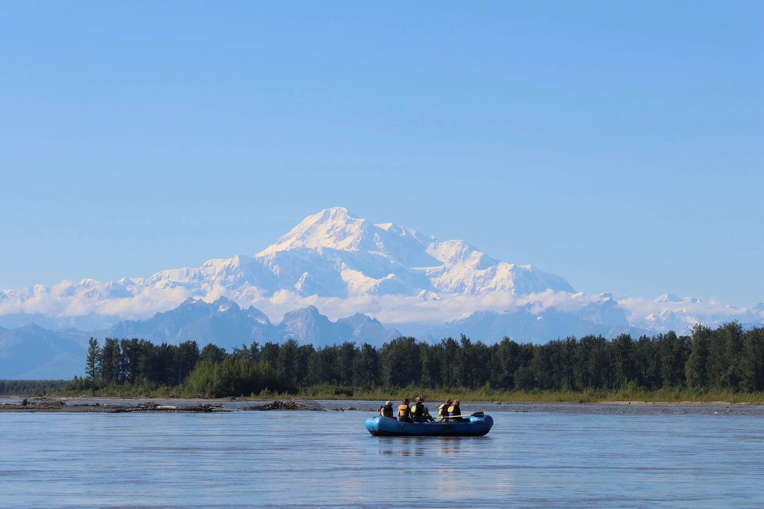 A group of people in a blue inflatable boat on a river, with a snow-capped mountain in the background, surrounded by forest and clear blue sky.