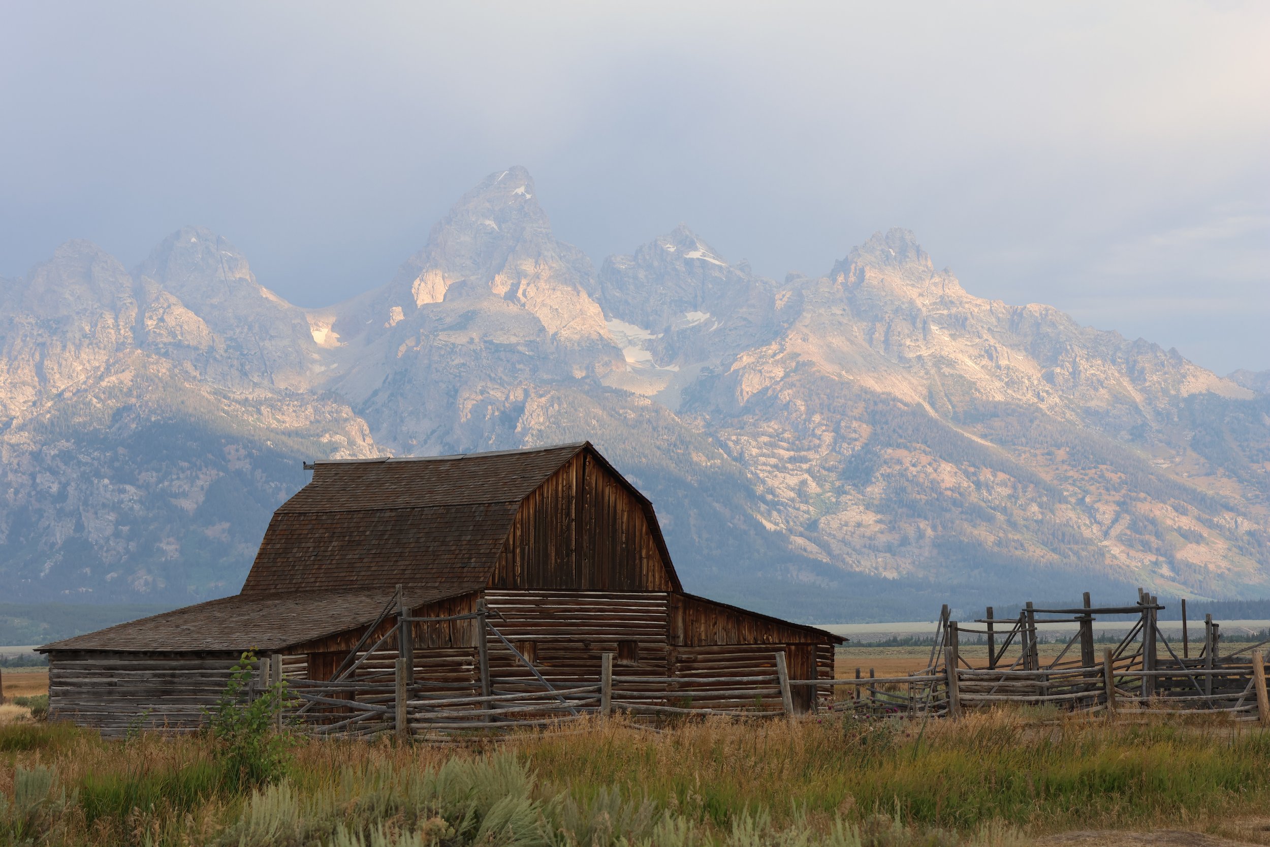 A rustic wooden barn with a mountain range featuring rocky peaks and patches of snow in the background, under a cloudy sky.