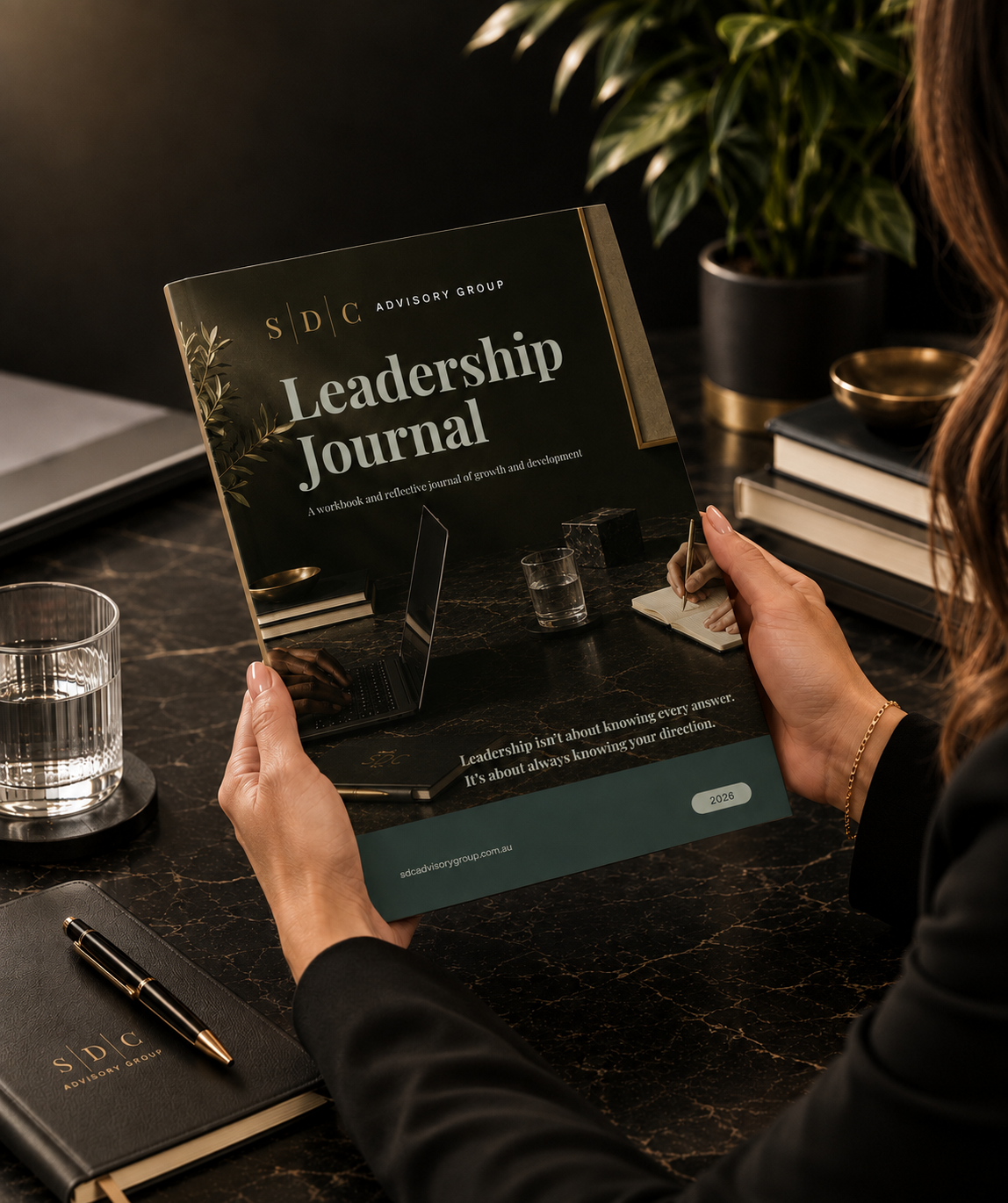 Person holding a Leadership Journal booklet at a desk with books, a glass of water, notebook, and pen.