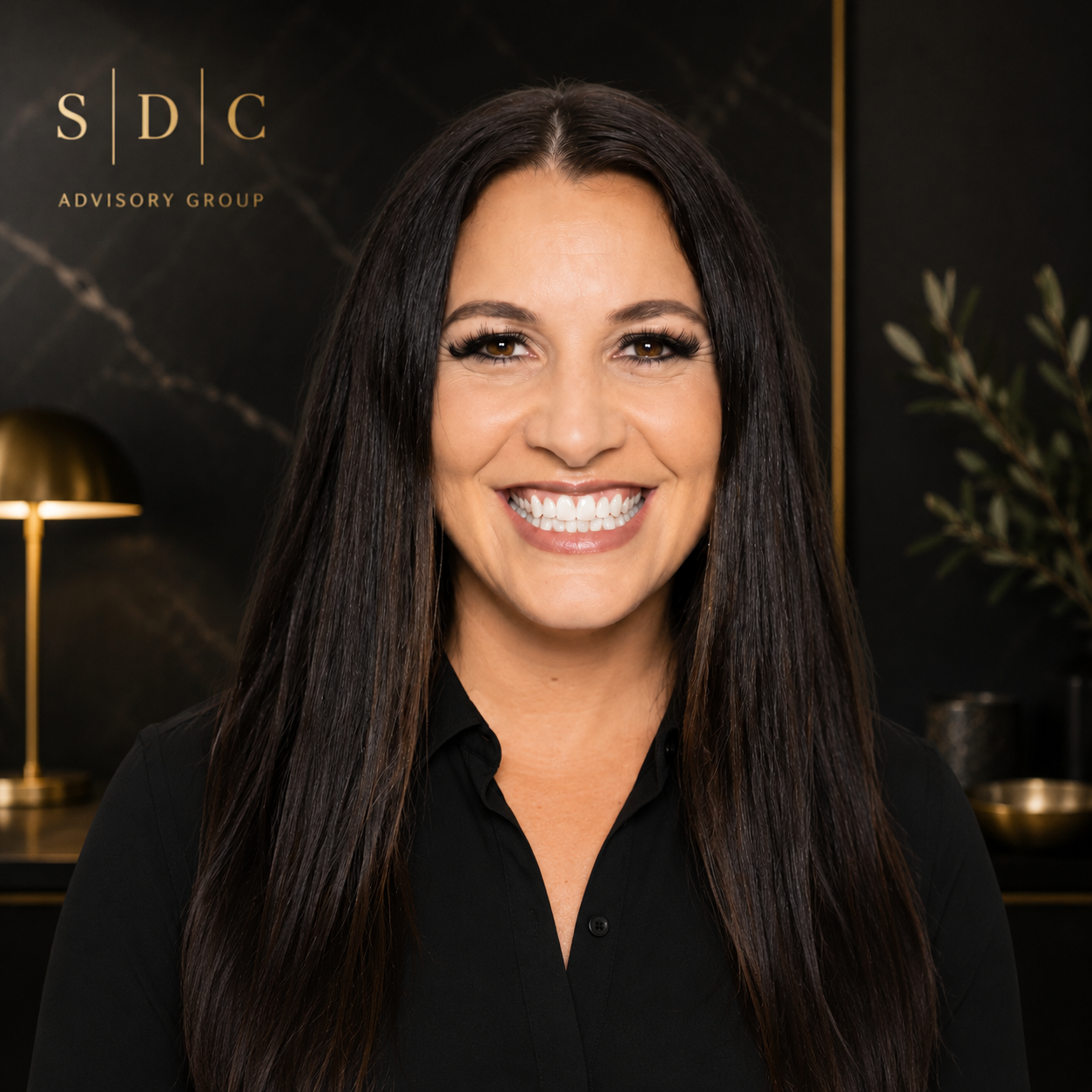 A woman smiling with long dark hair, wearing a black shirt, standing in a modern office with a black marble wall, gold accents, a lamp, and a plant in the background.