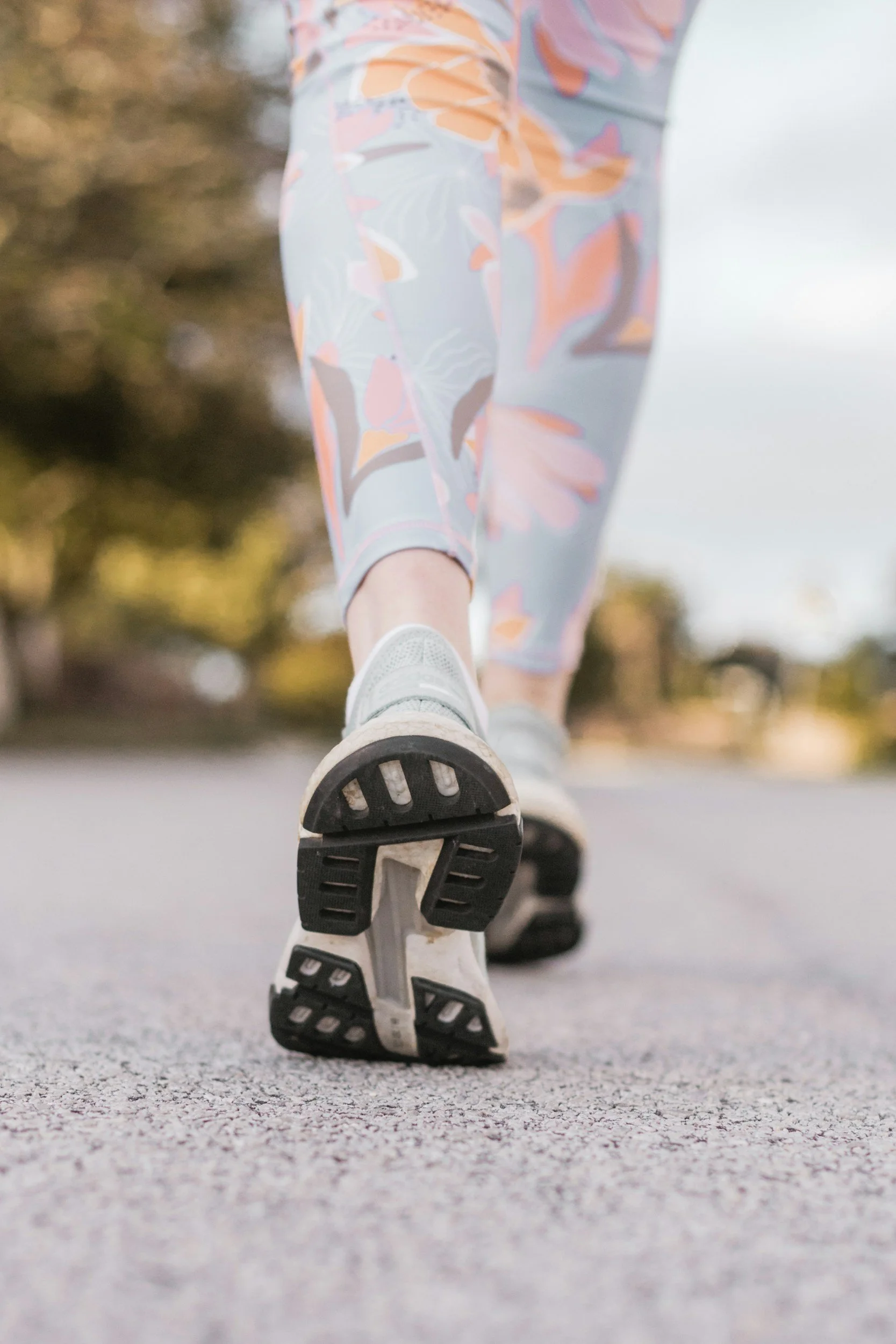 Close-up of a person's legs and feet wearing white sneakers and colorful patterned leggings, walking on a paved path in an outdoor setting with trees and blurred background.