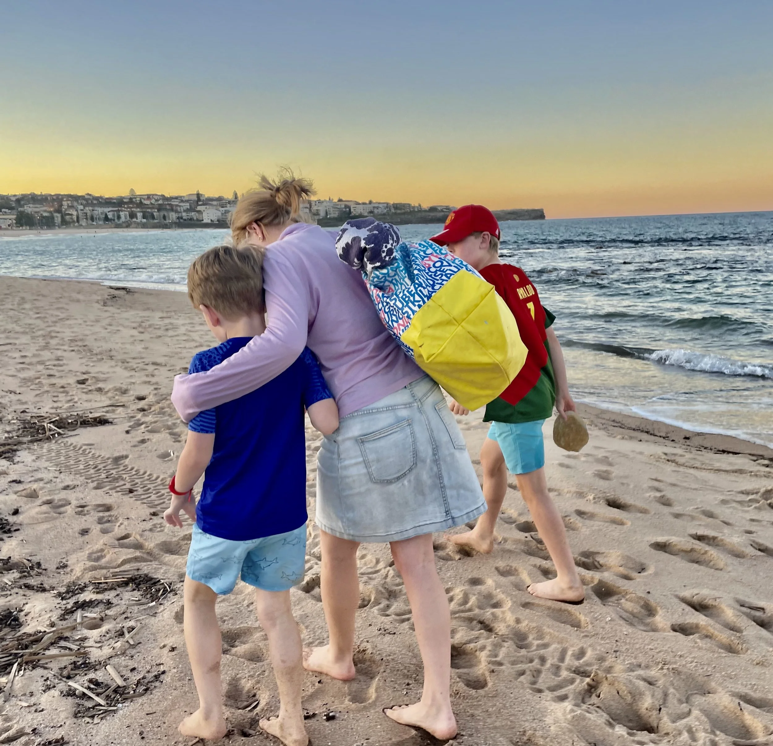 A woman and three children walking barefoot on a sandy beach at sunset, with a coastal town visible in the background.