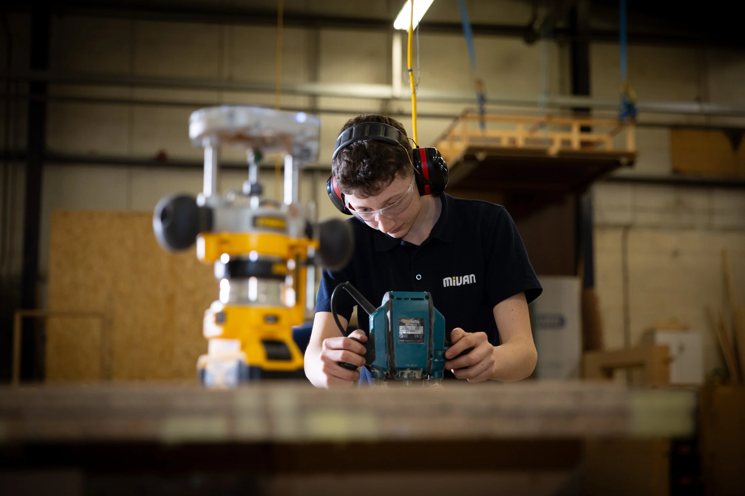 A young apprentice working in a woodworking shop, wearing safety glasses and ear protection, focused on using a power tool.