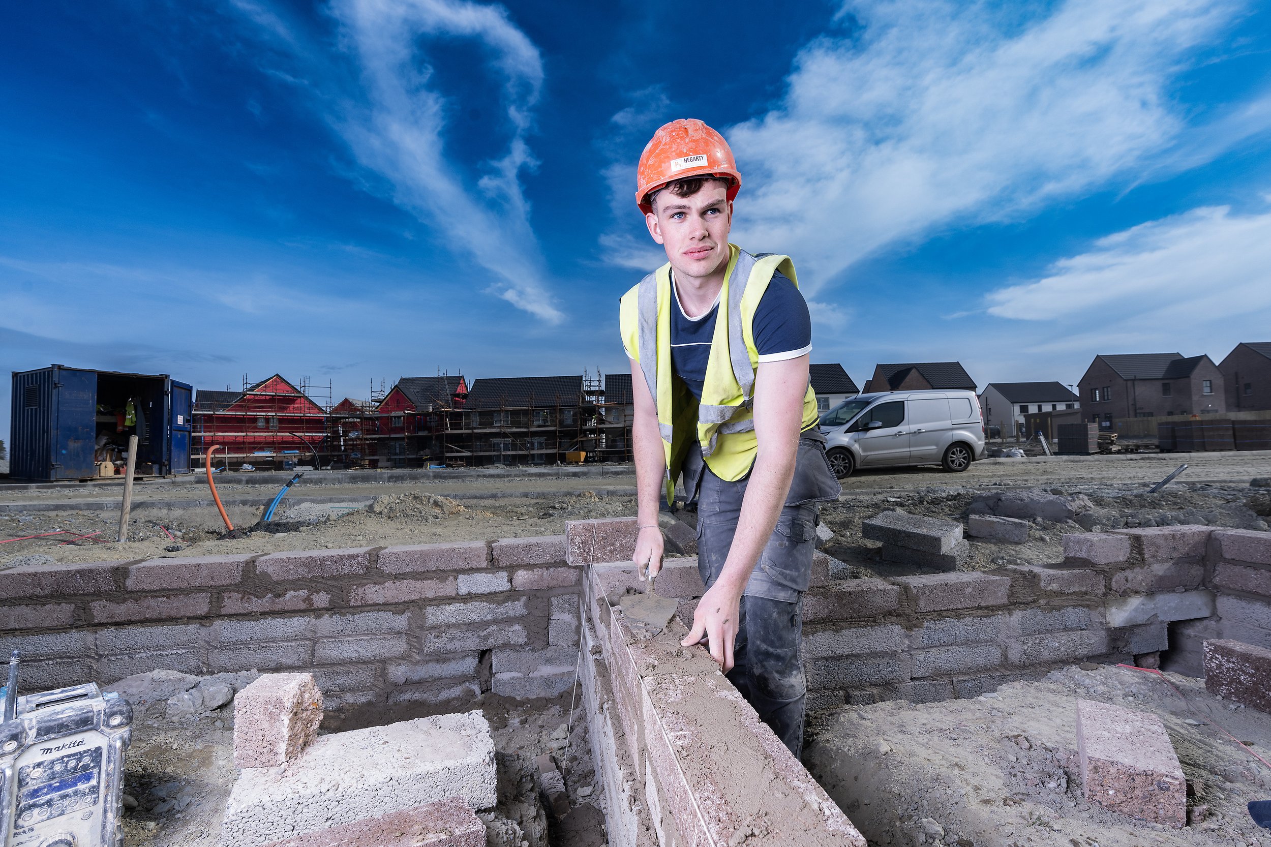 An apprentice construction worker with a red hard hat and yellow safety vest, laying bricks for a building foundation on a construction site. There are houses under construction and parked vehicles in the background under a blue sky.