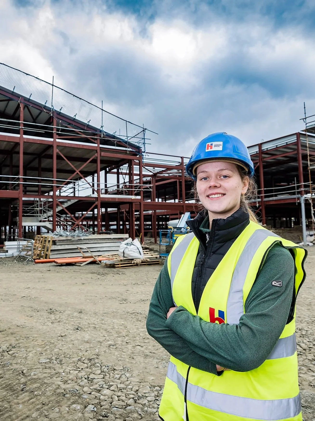 A young construction apprentice in a hard hat and high-visibility vest standing on a construction site in Northern Ireland with scaffolding and building materials in the background.