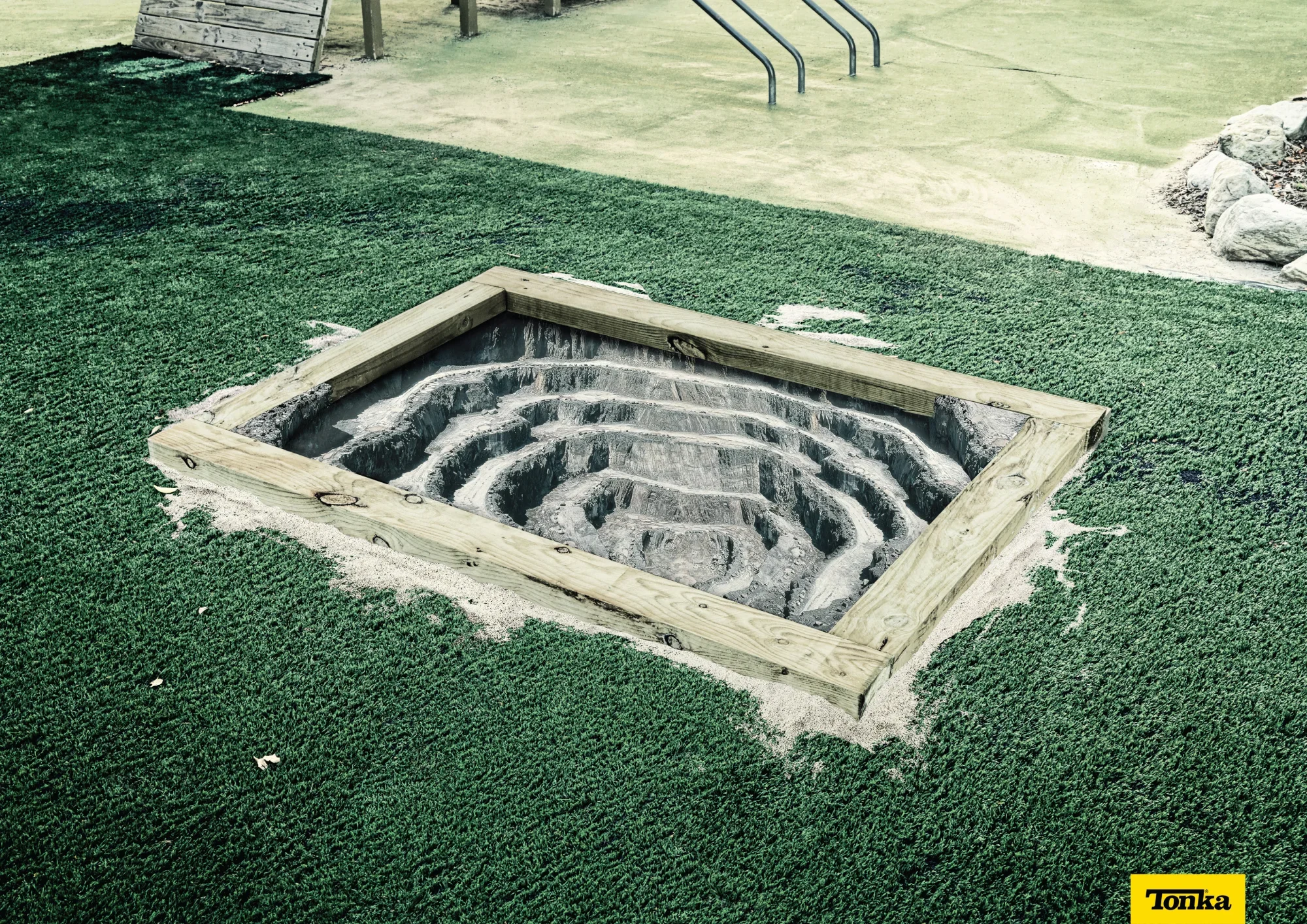 A square wooden frame surrounds a deep, spiral-shaped sand trap or hole in a miniature golf course with artificial green turf and a concrete surface in the background.