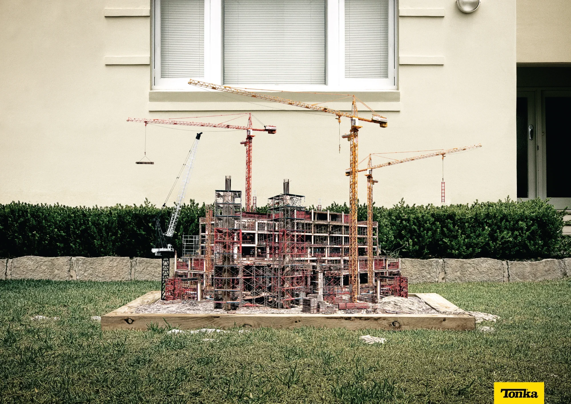 Miniature construction site model with multiple cranes, scaffolding, and building framework placed on a patch of grass in front of a beige house exterior with window.
