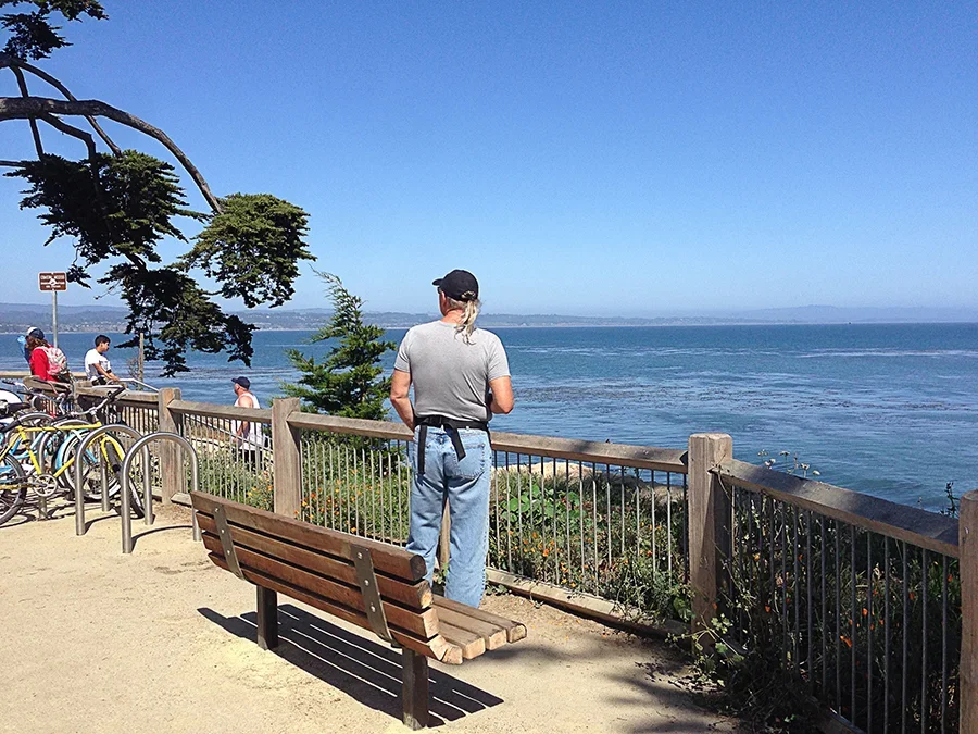 Person standing and looking at the ocean from a viewing area with a wooden bench and railing, bicycles parked nearby, and other visitors in the background on a sunny day.