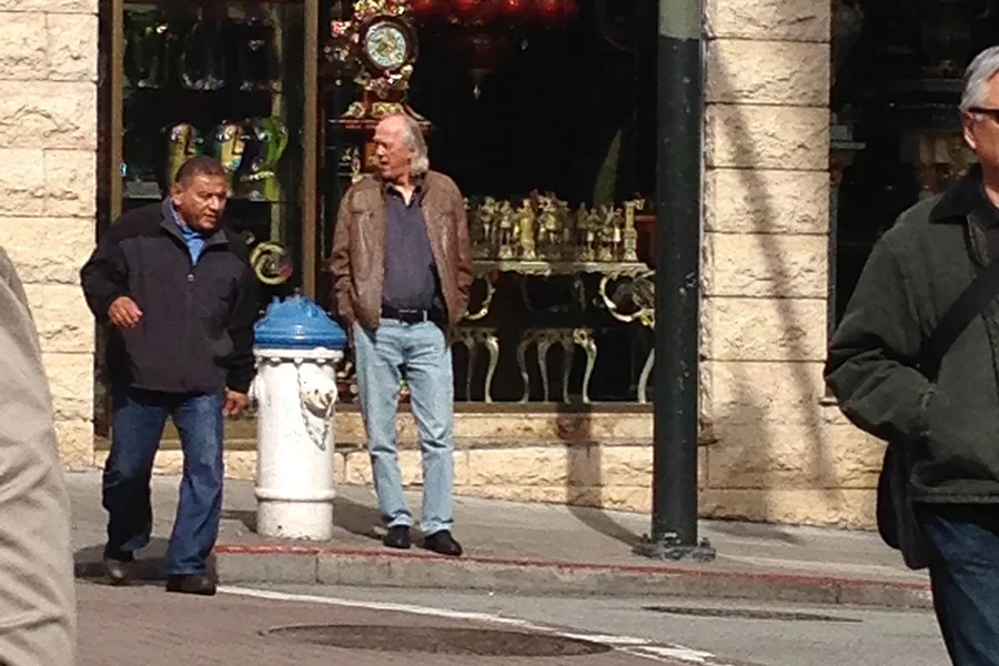 Street scene with three men standing near a shopwindow displaying clocks and antiques, with a trash can in the foreground.