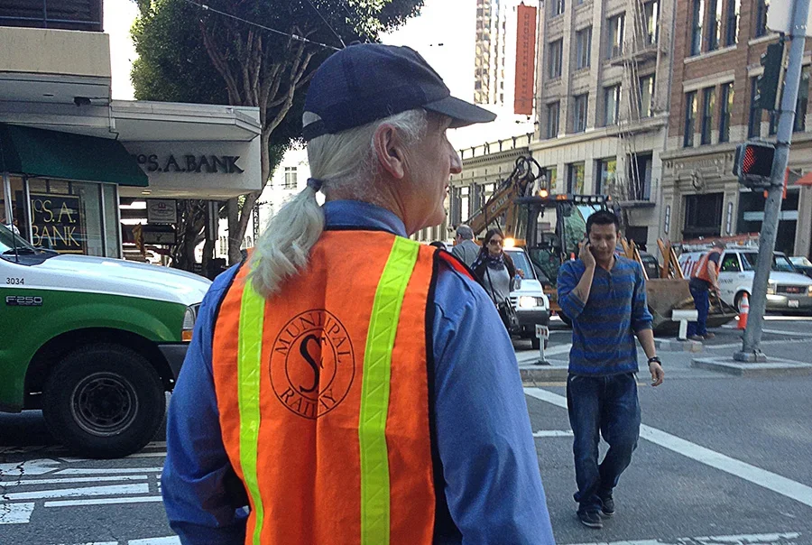 A woman wearing an orange safety vest with 'Municipal Worker' printed on the back, standing in a city street at an intersection, with pedestrians and vehicles around.