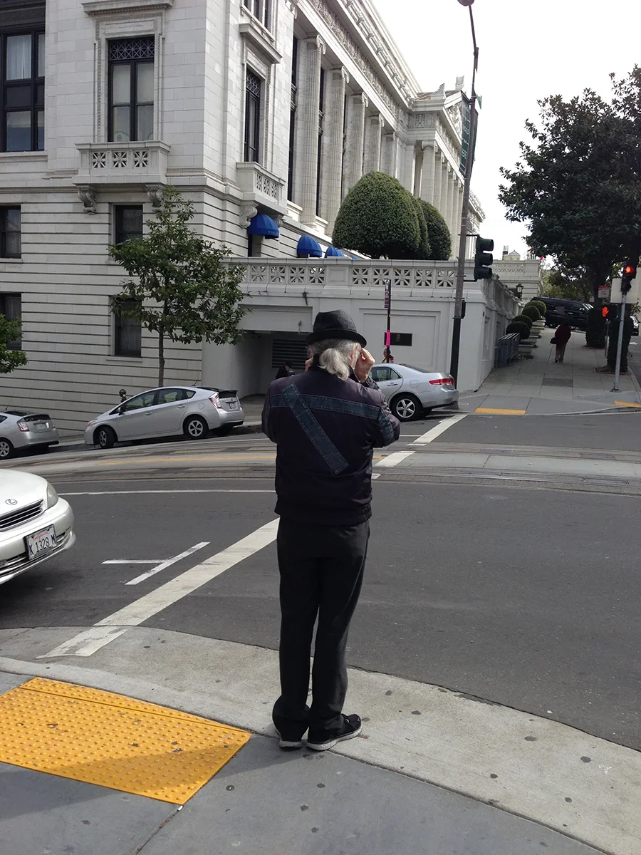 An older man with gray hair and a black hat stands on a city sidewalk, looking at his phone. He is wearing a dark jacket and black pants. Behind him is a street with parked cars, a large white historic building with architectural details, and some trees.