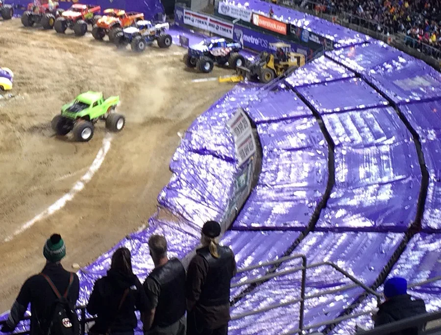 Monster trucks racing on a dirt track, with a spectator area and a few people watching from the bleachers.