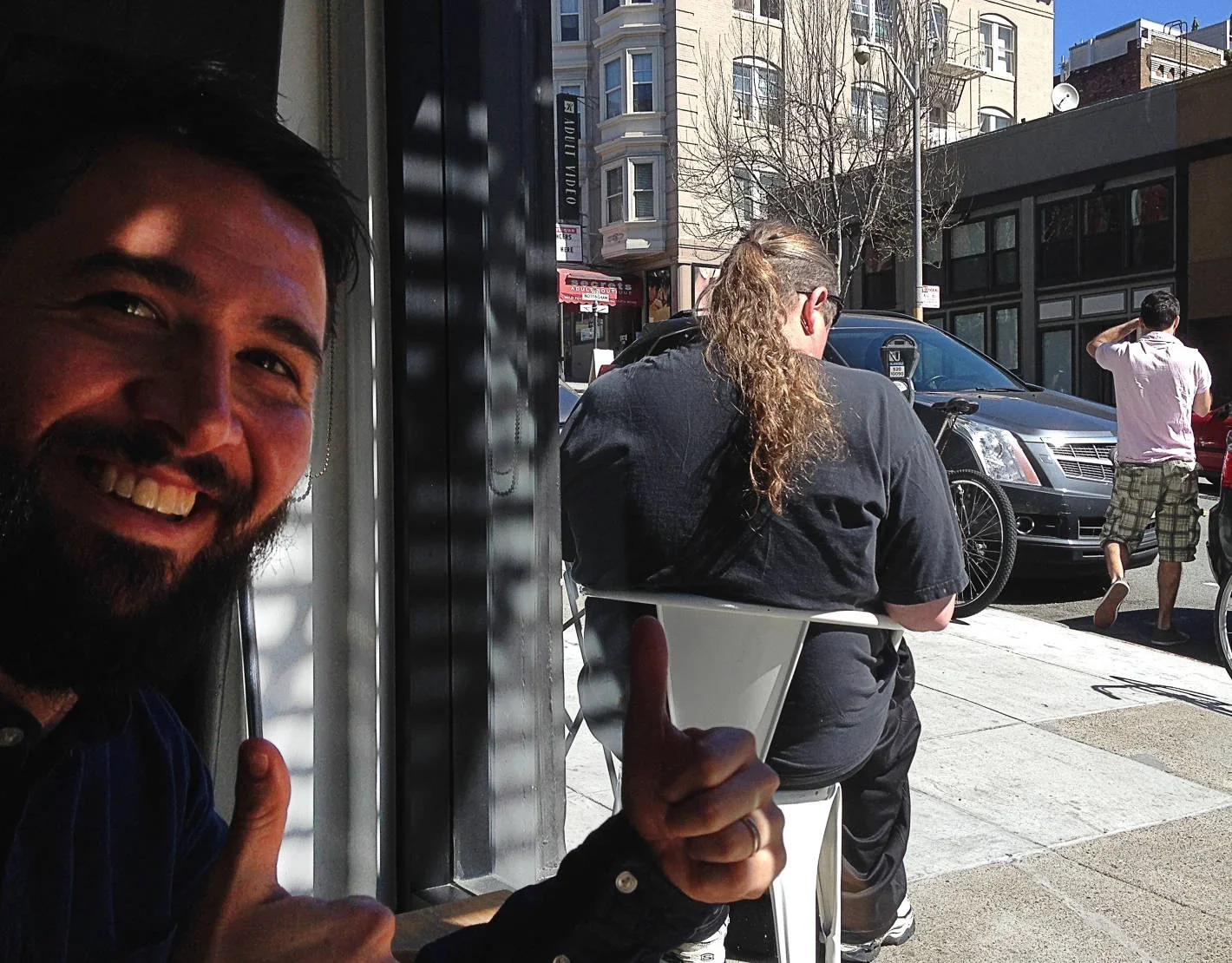 Smiling man with a beard giving a thumbs-up inside a cafe, with a woman sitting outside on a sidewalk at a table, and pedestrians walking by in an urban setting with buildings and cars.