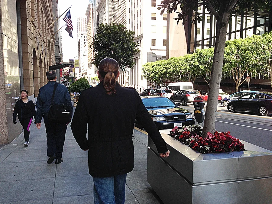 A woman with brown hair tied in a ponytail, dressed in a black coat and blue jeans, walking on a city sidewalk, with a flower planter containing red and white flowers and a small tree nearby.