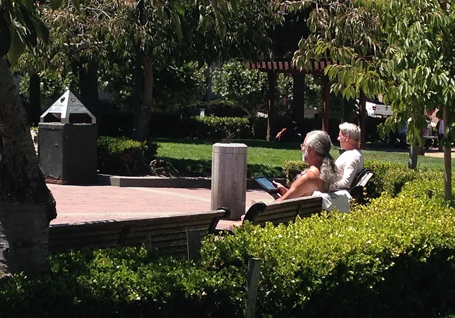 Two elderly people sitting on a park bench under shade trees, with one reading a tablet and the other holding a book, and greenery surrounding them.