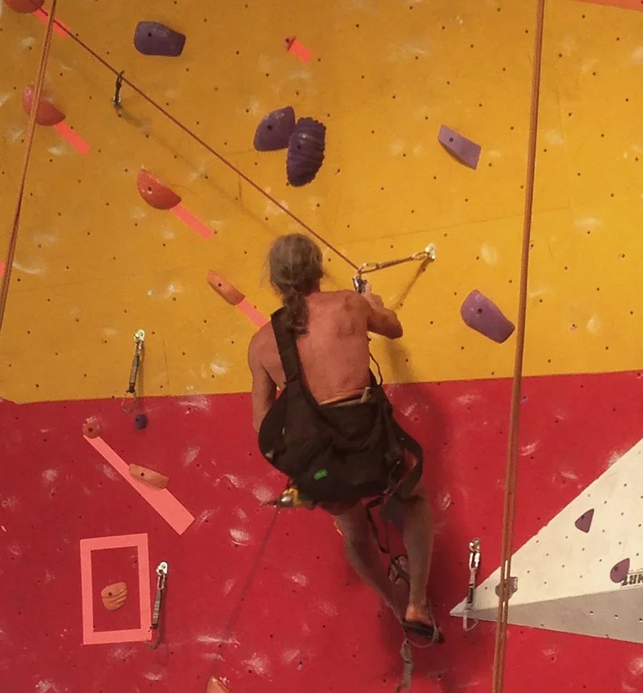 A man with long hair rock climbing on an indoor climbing wall, wearing a harness and using a belay device connected to a safety rope.
