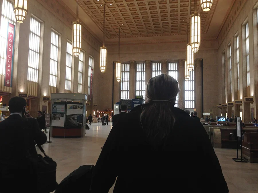 Interior of a busy train station with high ceilings, large windows, and hanging light fixtures. Several travelers are visible, including a woman with long hair in the foreground and others with luggage. Ticketing and information signs are present.