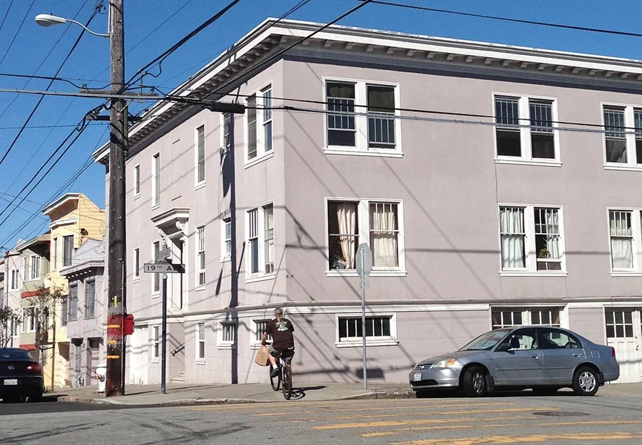A person riding a bicycle on a city street corner with a silver sedan parked nearby and a pale gray building in the background.