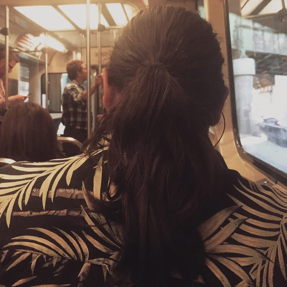 Person with dark hair sitting on a bus, wearing a black and white patterned top, looking out the window. In the background, there's a woman standing and speaking, and another person using a phone.