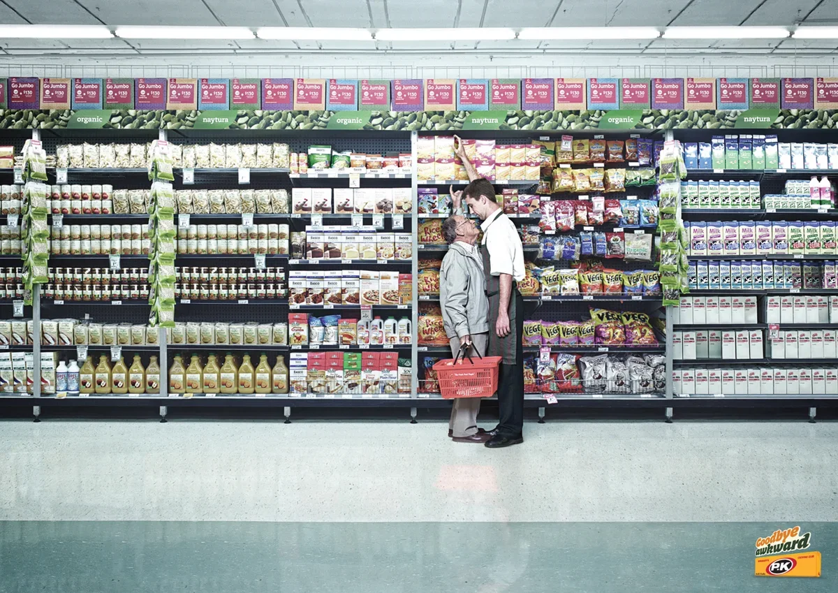 An elderly woman and a young man are standing closely together in an aisle of a grocery store, looking at each other. The woman is holding a red shopping basket, and the man is reaching up to a shelf filled with various snack items. The store shelves are stocked with cereal, snacks, and other grocery items, with a banner above advertising organic products.