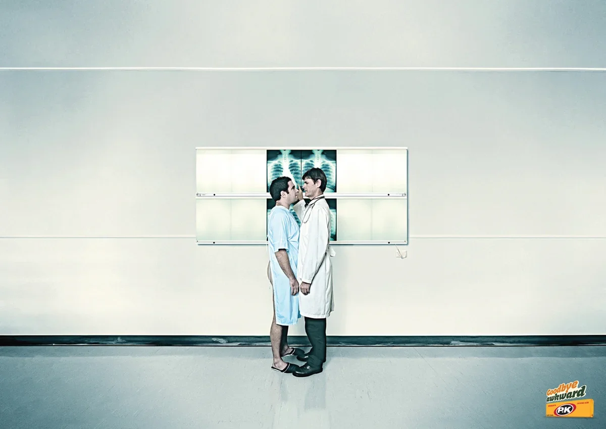 Two men, one in a hospital gown and the other in a white lab coat, stand facing each other in a hospital corridor with x-ray images on the wall behind them.