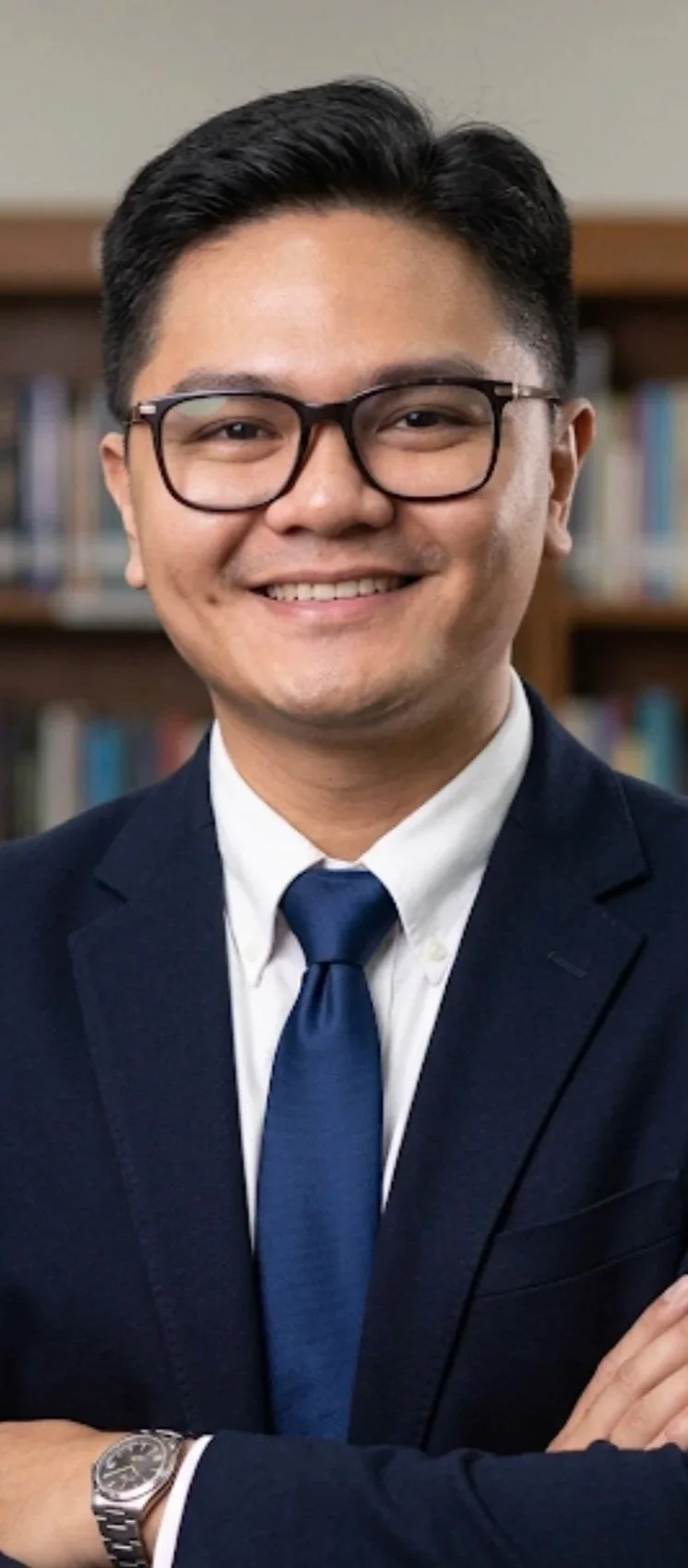 Headshot of a smiling young man in a suit and tie, wearing glasses, with a background of bookshelves.