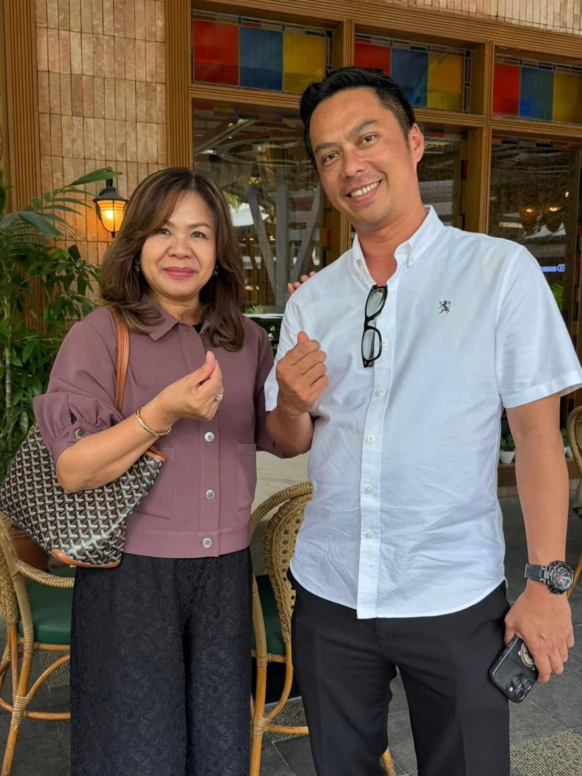 A woman and a man standing close together, both smiling and making a fist with their right hands. They are outside a restaurant or café with wicker chairs and plants in the background.