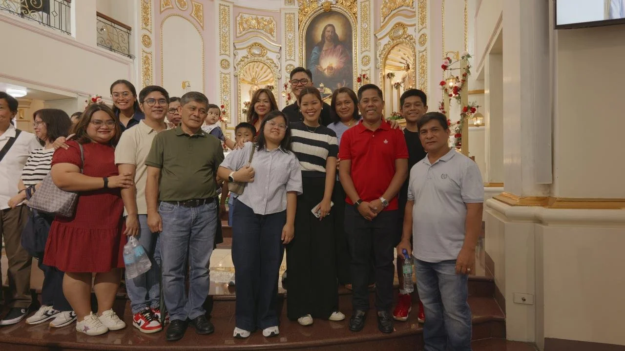 Group of people gathered inside a church, standing on steps in front of an ornate altar with religious iconography and a painting of Jesus Christ.