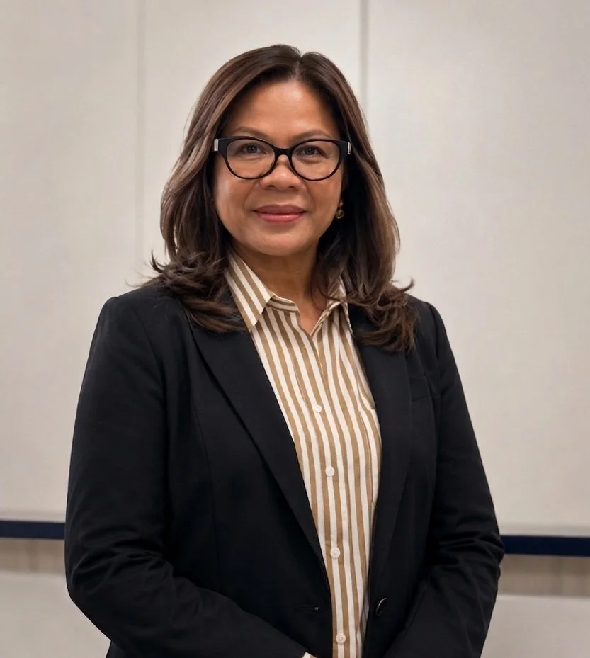 A woman wearing glasses, a black blazer, and a beige and white striped shirt, standing in front of a plain wall.