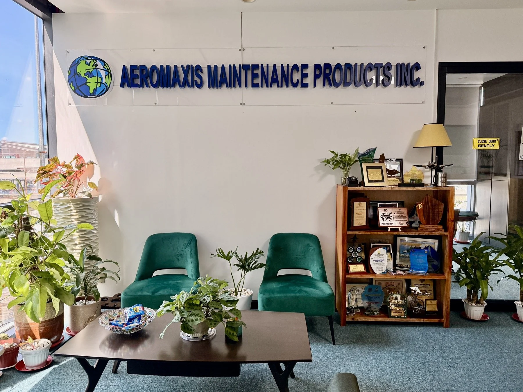 Office reception area with greenery, two green chairs, a wooden shelf with awards and decorations, and a sign on the wall that reads 'AEROMAXIS MAINTENANCE PRODUCTS INC.' with a globe logo.