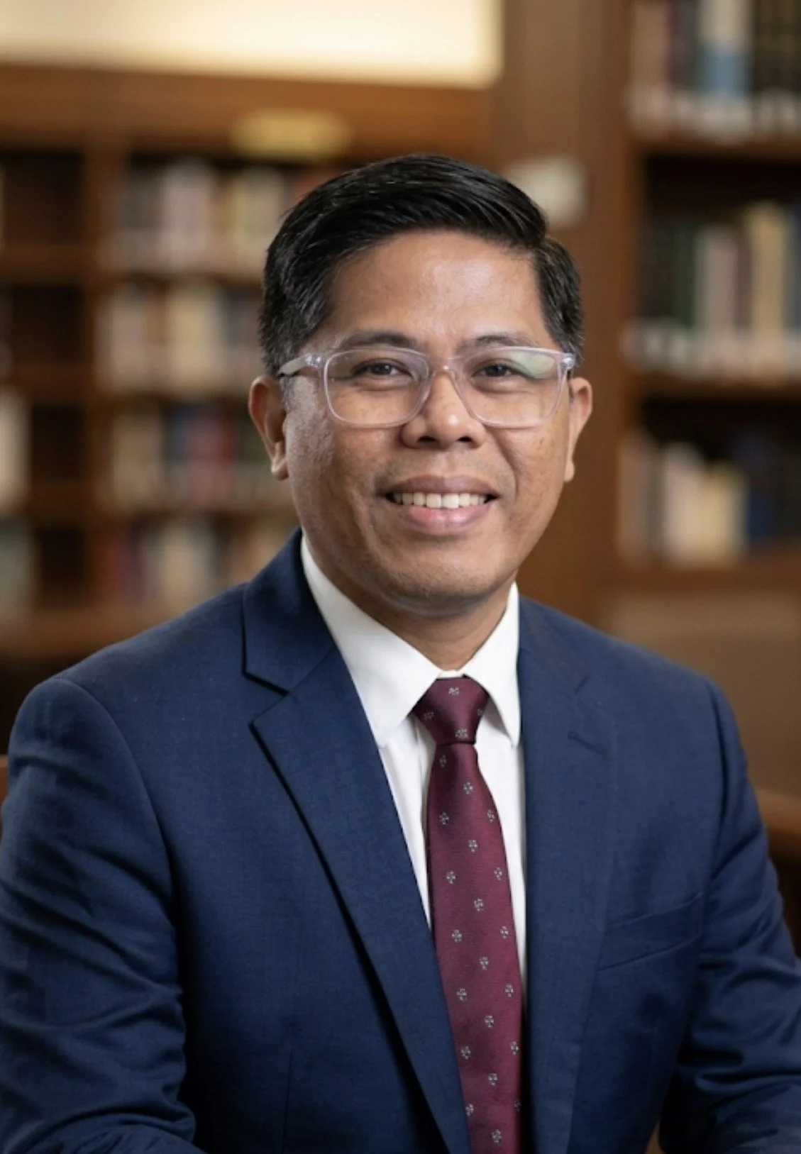 Portrait of a man in a suit with glasses, smiling, sitting in front of a bookshelf in a library or office setting.