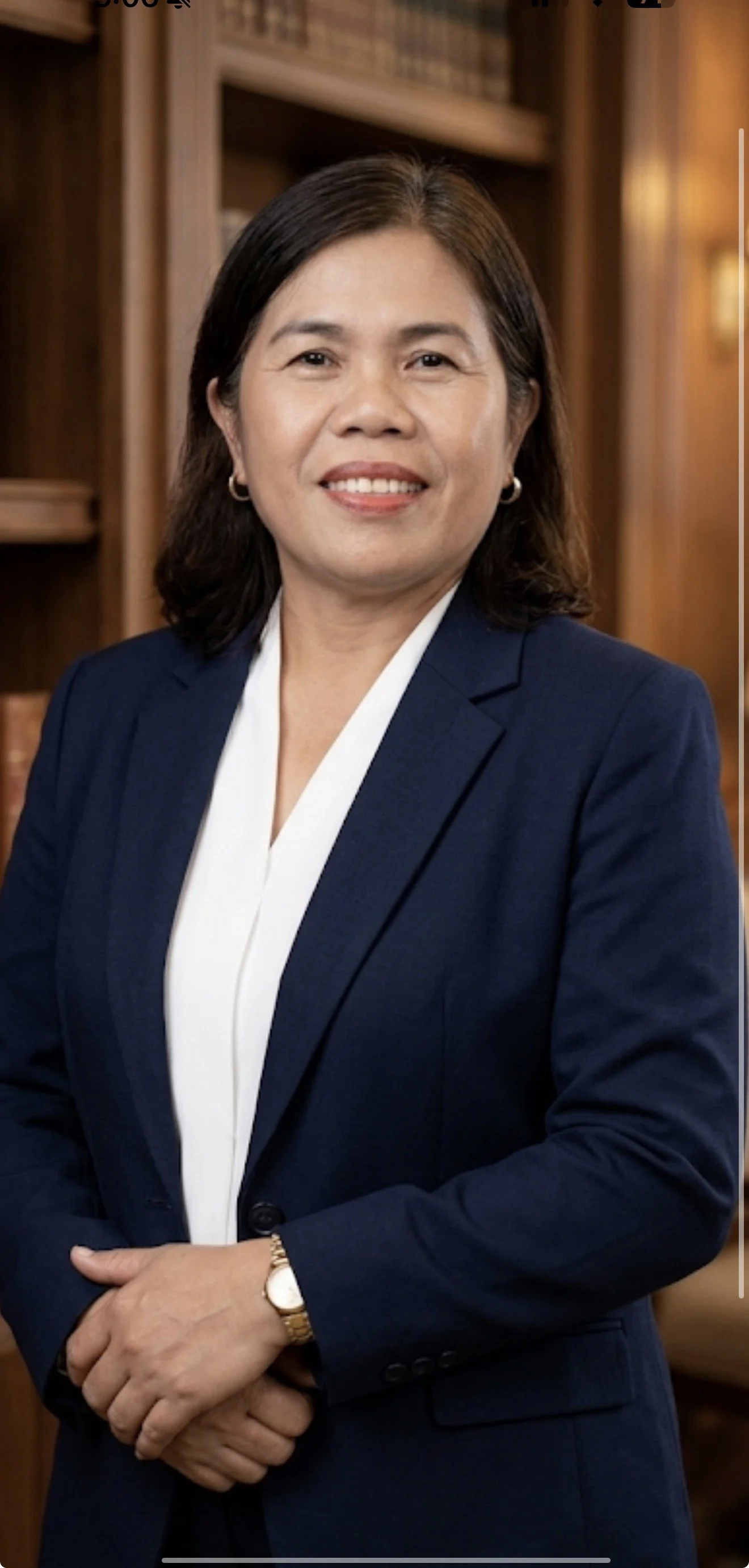 A professional woman with shoulder-length dark hair, wearing a navy blazer and white blouse, standing in a wood-paneled room with bookshelves in the background, smiling at the camera.