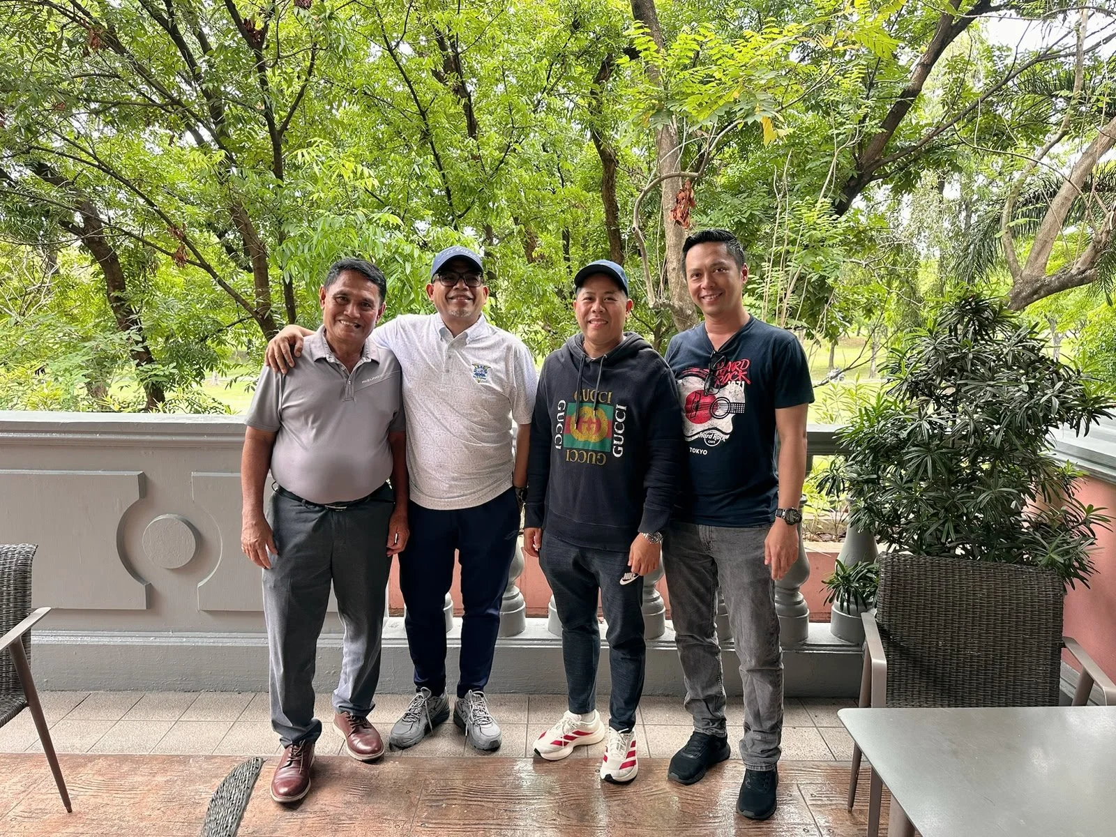 Four men standing outdoors on a balcony, surrounded by green trees.