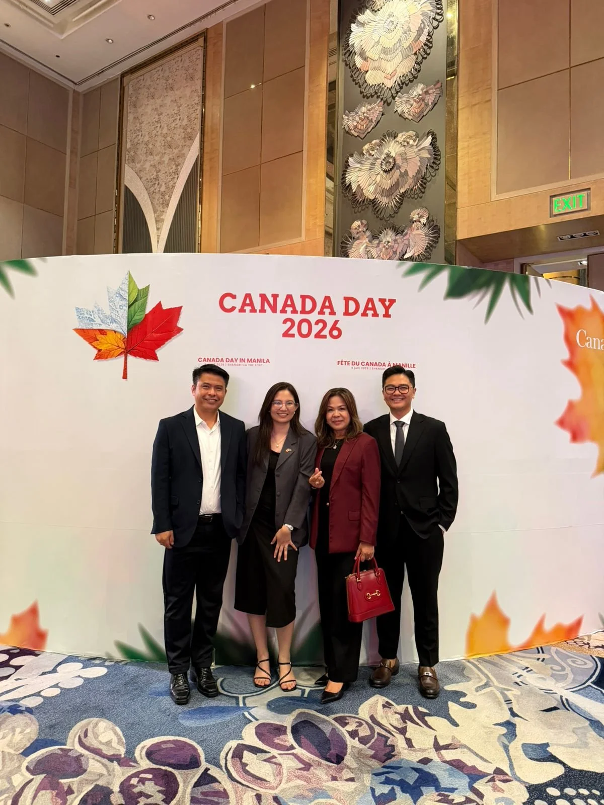 Four people dressed in formal attire standing in front of a backdrop that reads 'Canada Day 2026' with a maple leaf graphic. The venue is decorated with elegant wall art and a patterned carpet.