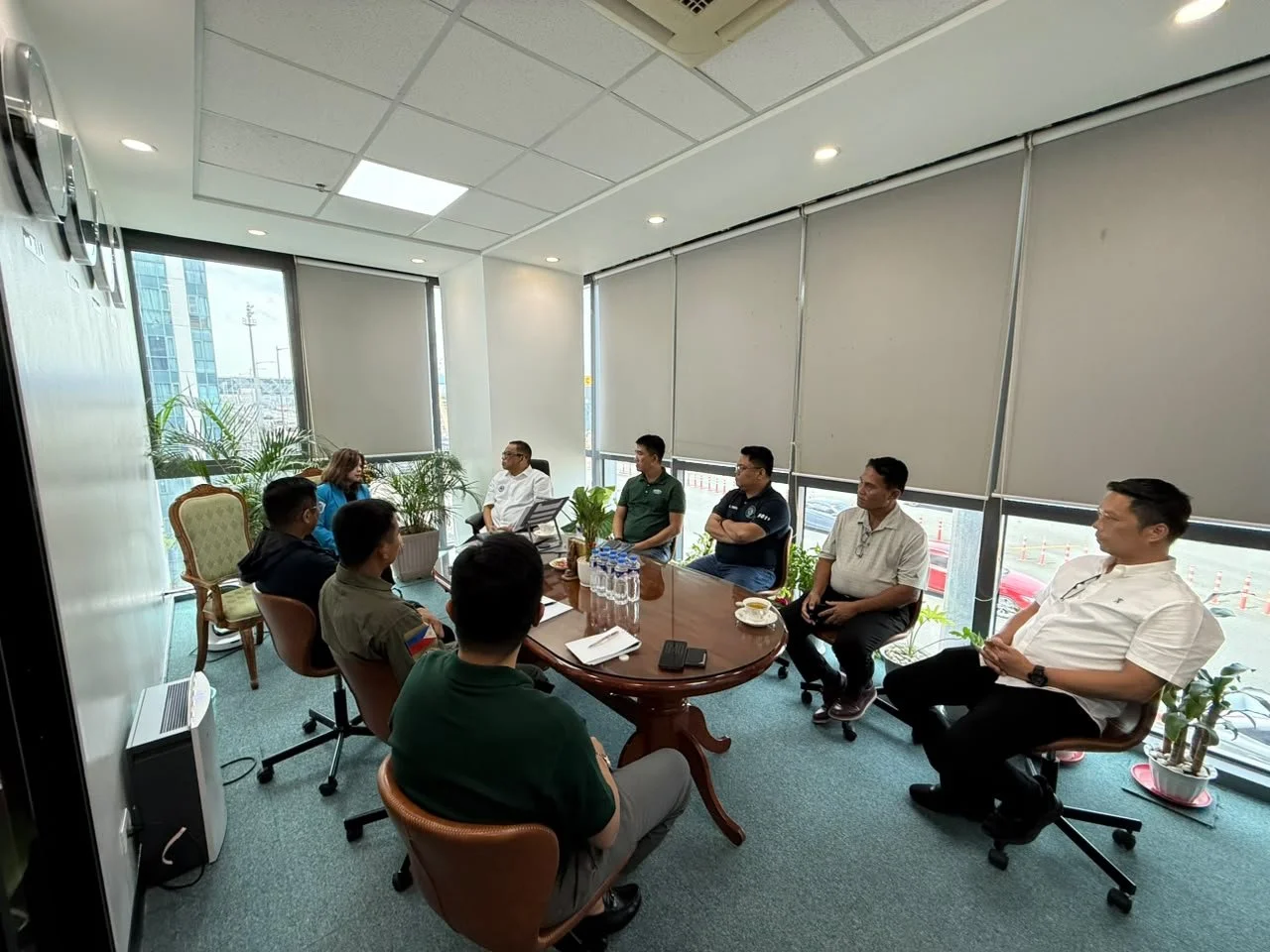 A group of people sitting around a conference table in a modern office with large windows, plants, and closed blinds.