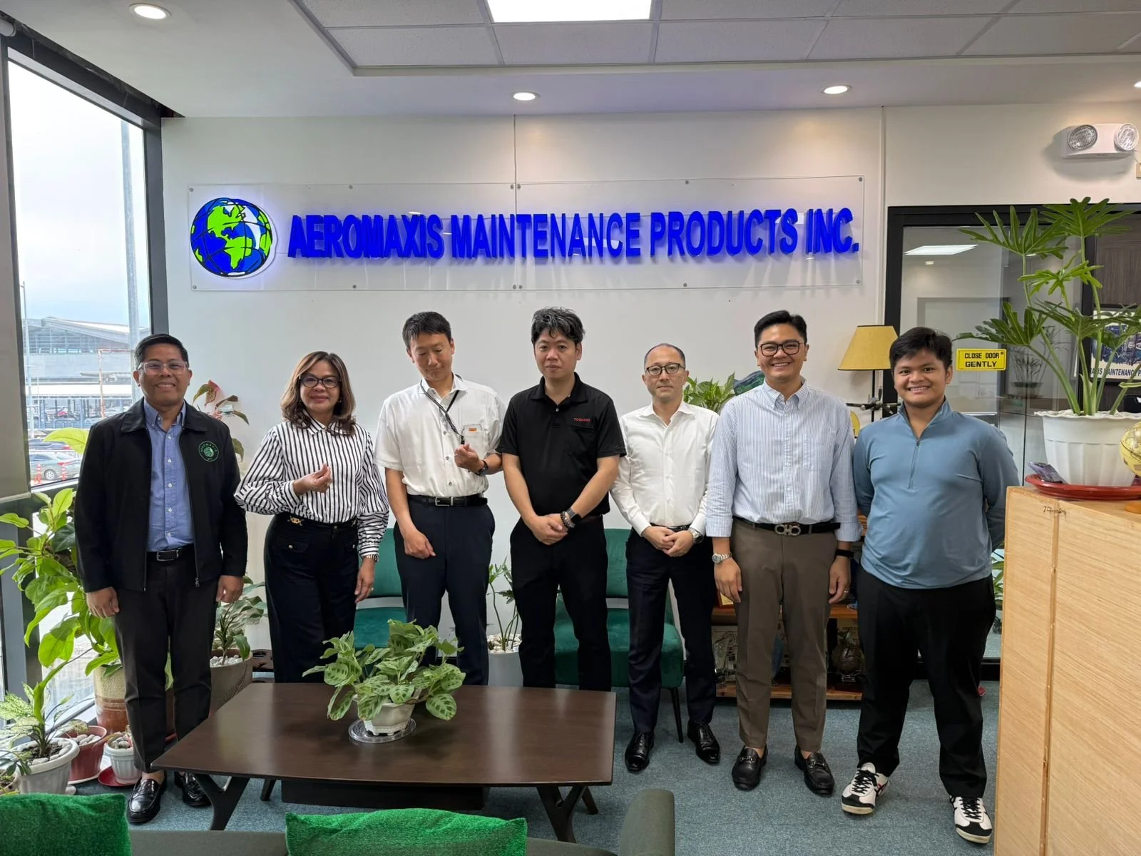 A group of seven people standing in an office with plants, in front of a sign that reads 'AEROMAXIS MAINTENANCE PRODUCTS INC.' The group appears to be colleagues or team members posing for a photo.