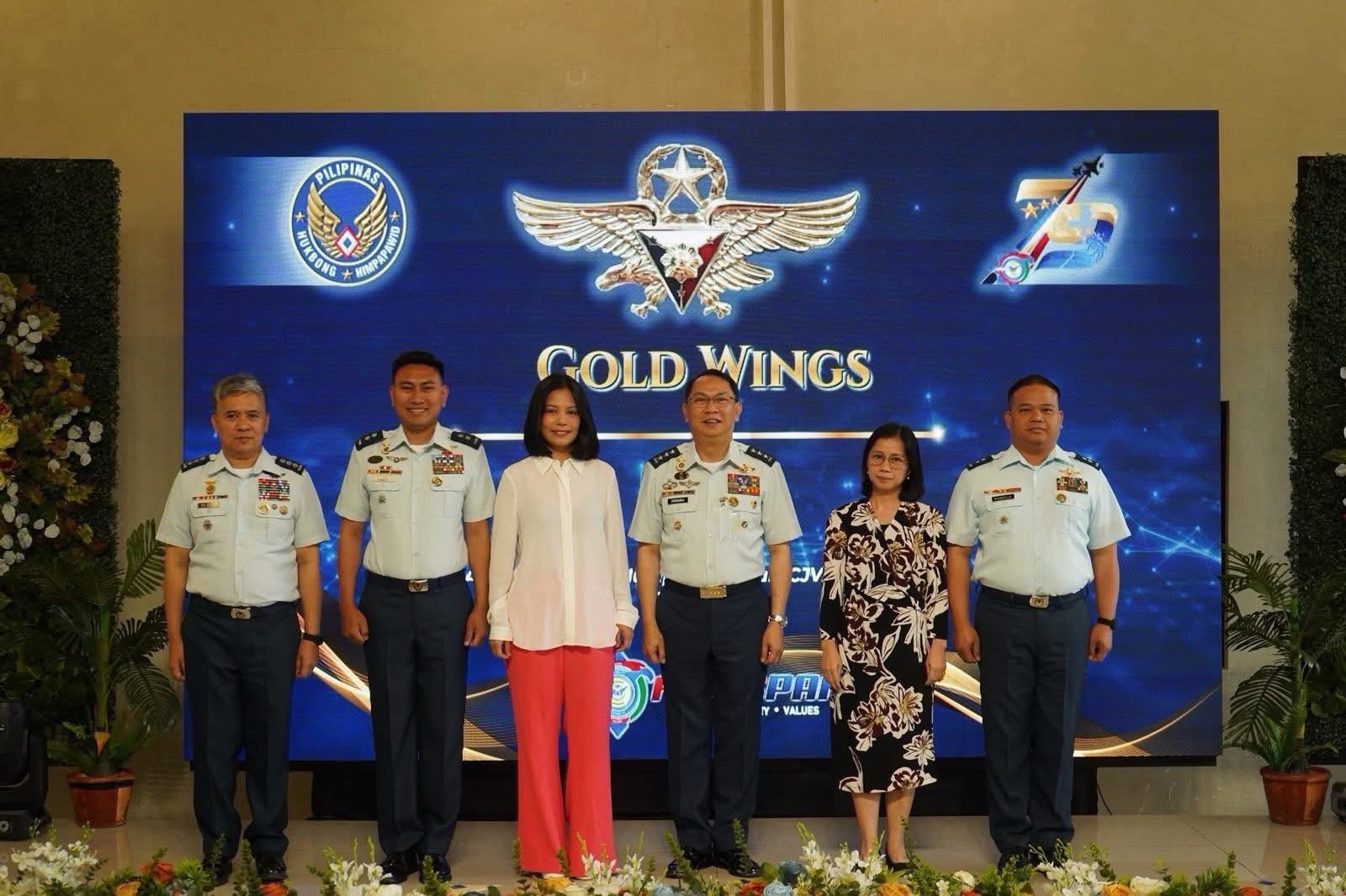 Group of six people, including five men in uniform and one woman in civilian clothing, standing in front of a large blue screen with military logos and the text 'Gold Wings'. Flowers are at their feet.