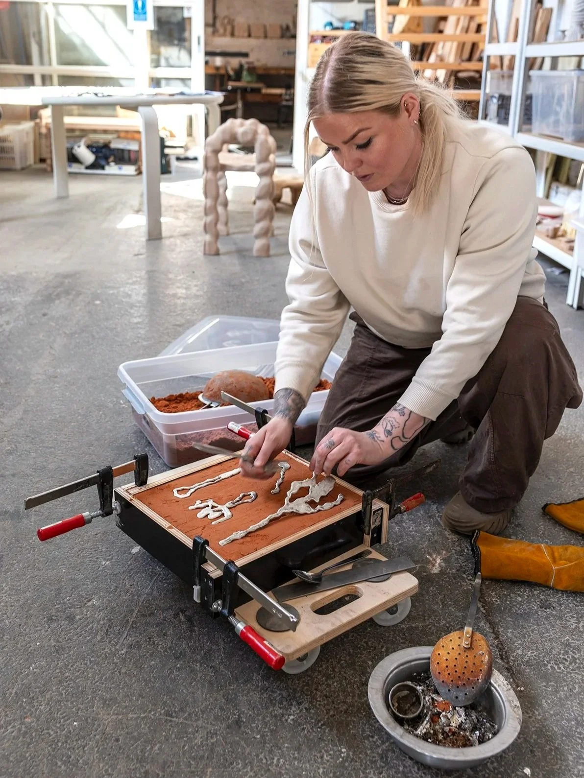 A woman with tattoos, blonde hair, and wearing a cream-colored sweater and brown pants kneels on the floor, customizing a small wooden tray with white glue, in a workshop with woodworking tools and furniture. There is a table, shelves, and a decorative sculpture in the background.