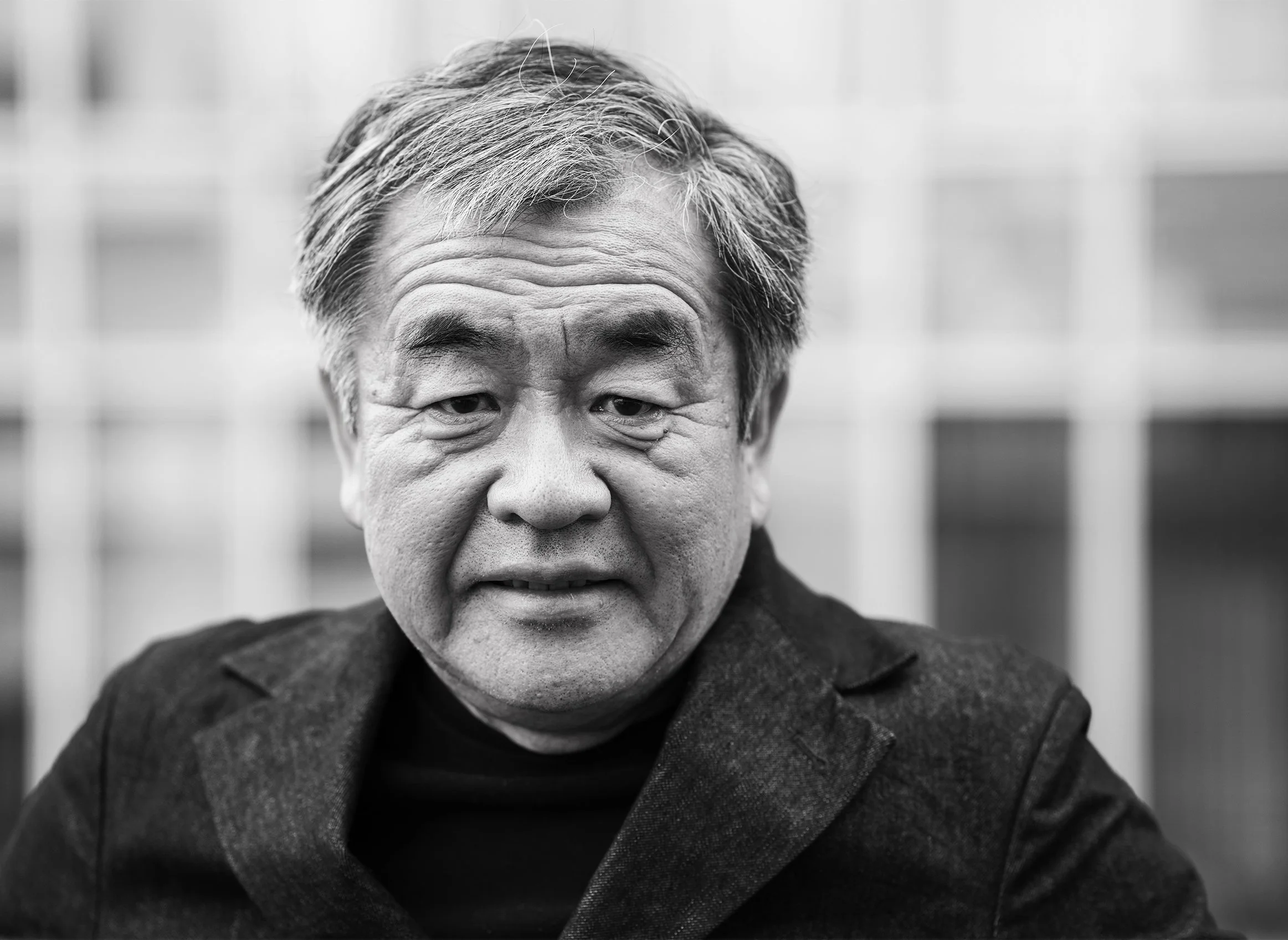 Close-up black and white photograph of an older Asian man with short hair and expressive facial lines, wearing a dark coat, with a blurred background.