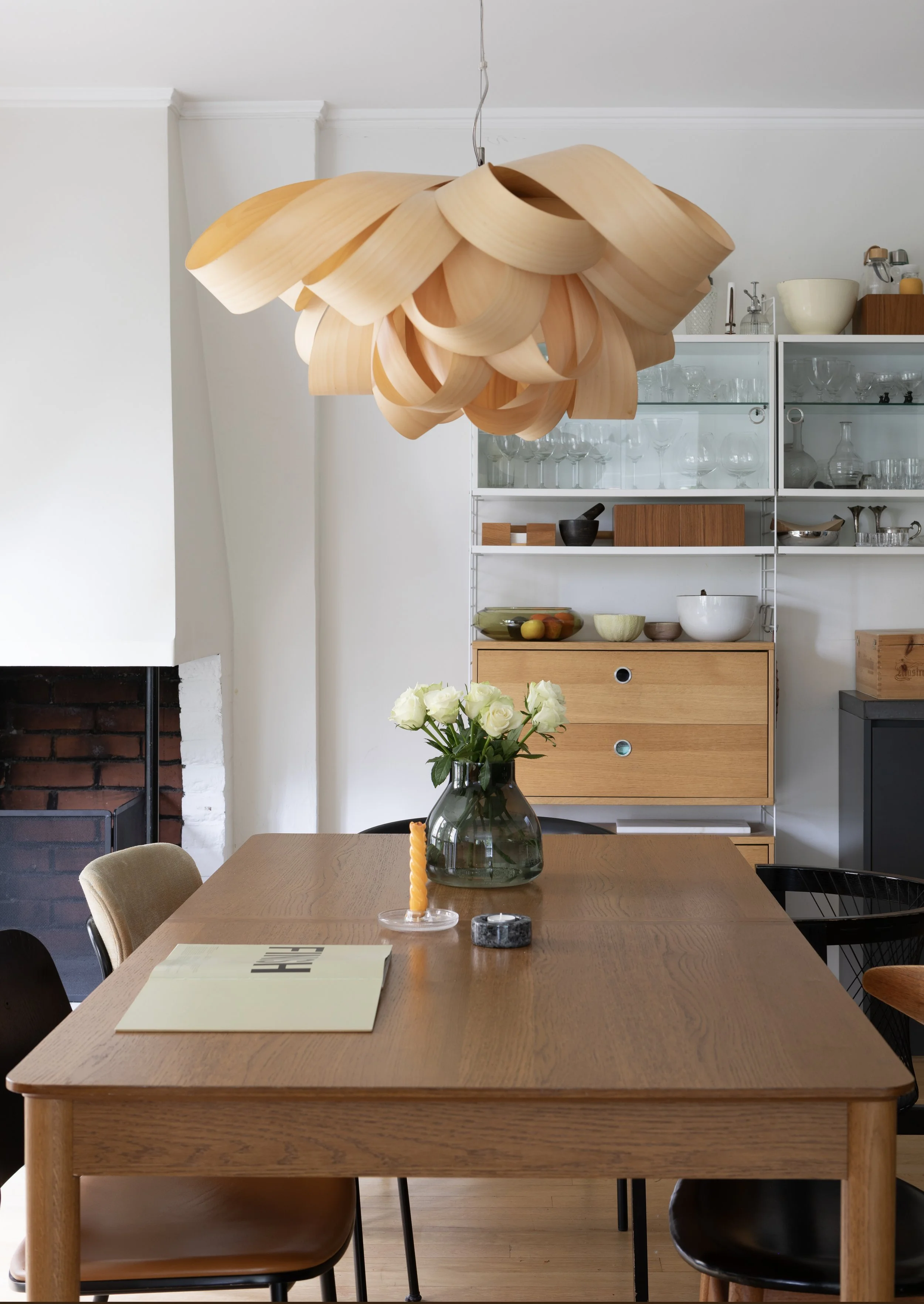 A modern dining room with a large wooden table, surrounded by chairs and a vase of white roses. A unique wooden pendant light hangs from the ceiling, and a white shelving unit with glassware and decorative items is in the background.