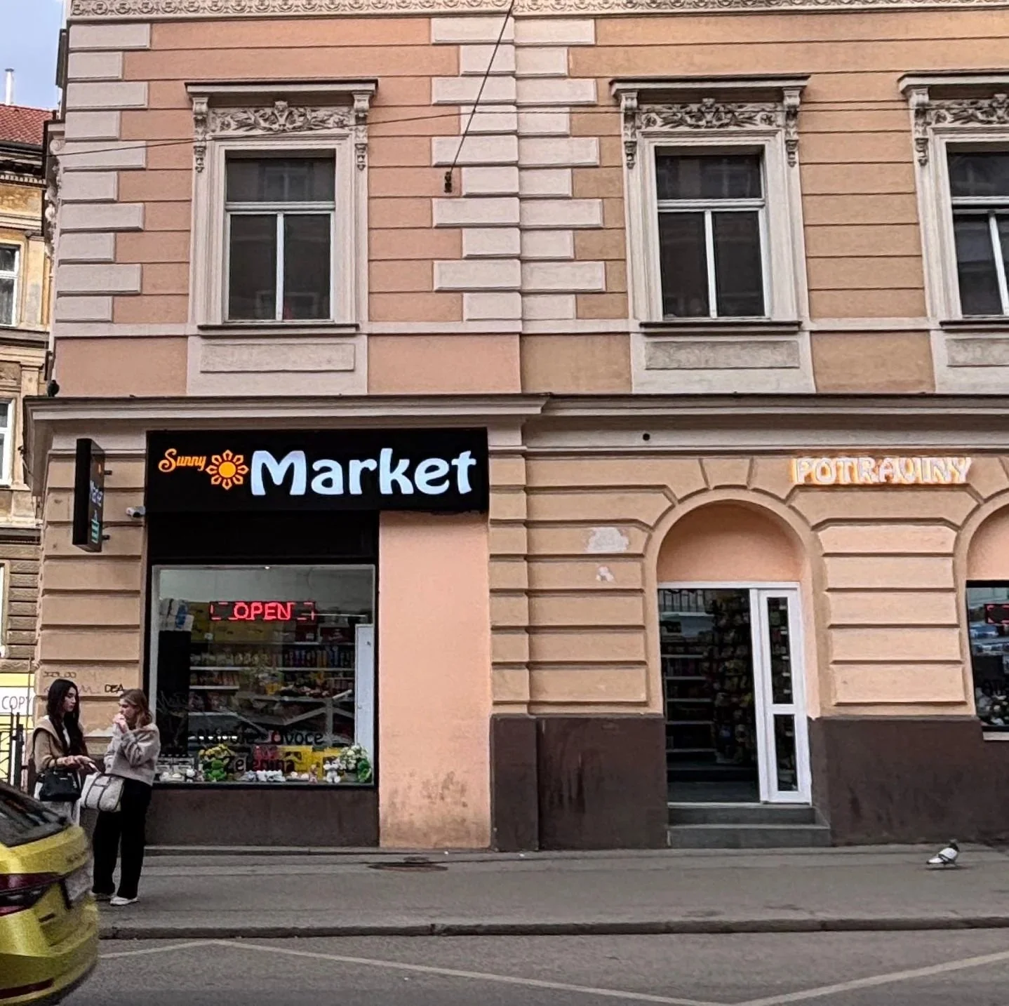 Street view of a corner store with signs for Sunny Market and Potraviny, a small grocery store with an open sign in the window, two women talking on the sidewalk, a yellow car parked in front, and a pigeon on the curb.