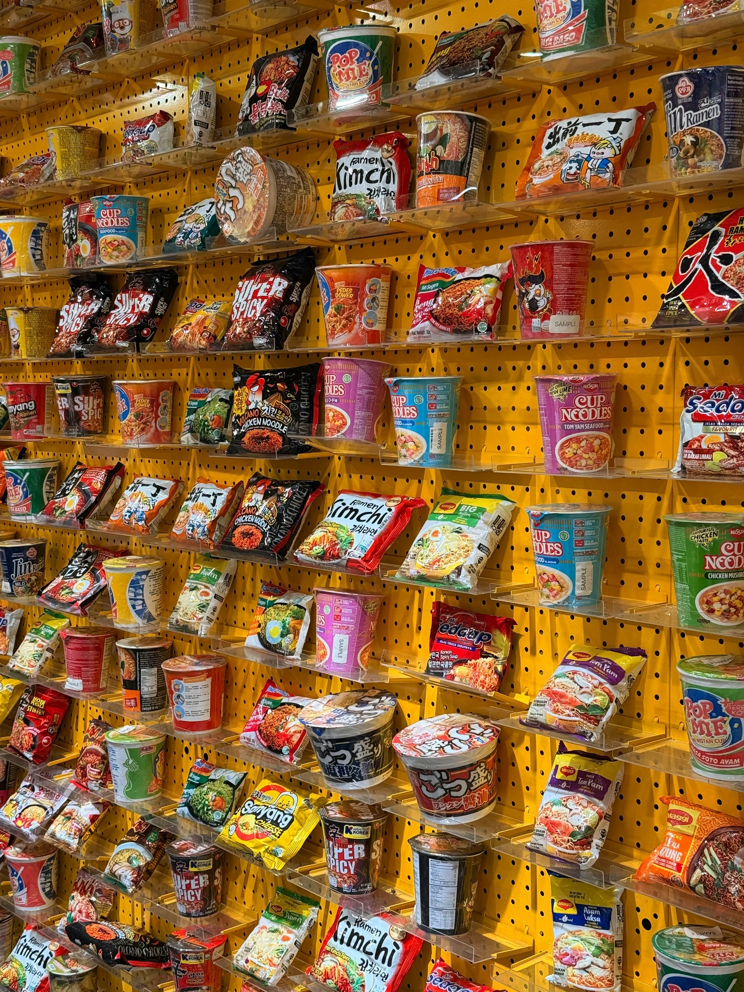 A store shelf filled with various instant noodle cups and packets in different colors and brands, arranged on yellow-colored pegboard.