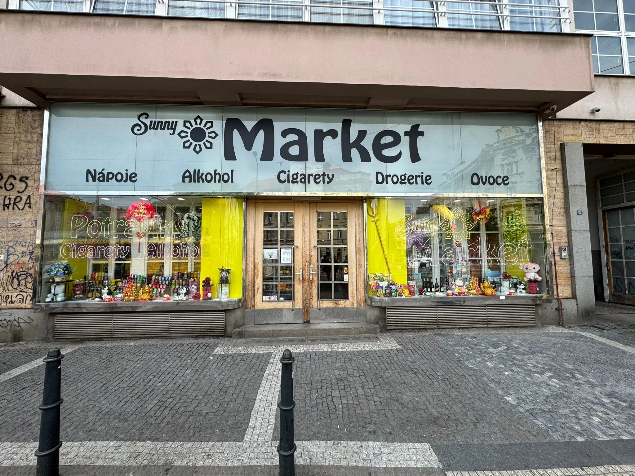 A storefront with a sign reading 'Sunny Market' and smaller words underneath listing beverages, alcohol, cigarettes, toiletries, and fruits. The store has a large window display with various items inside.