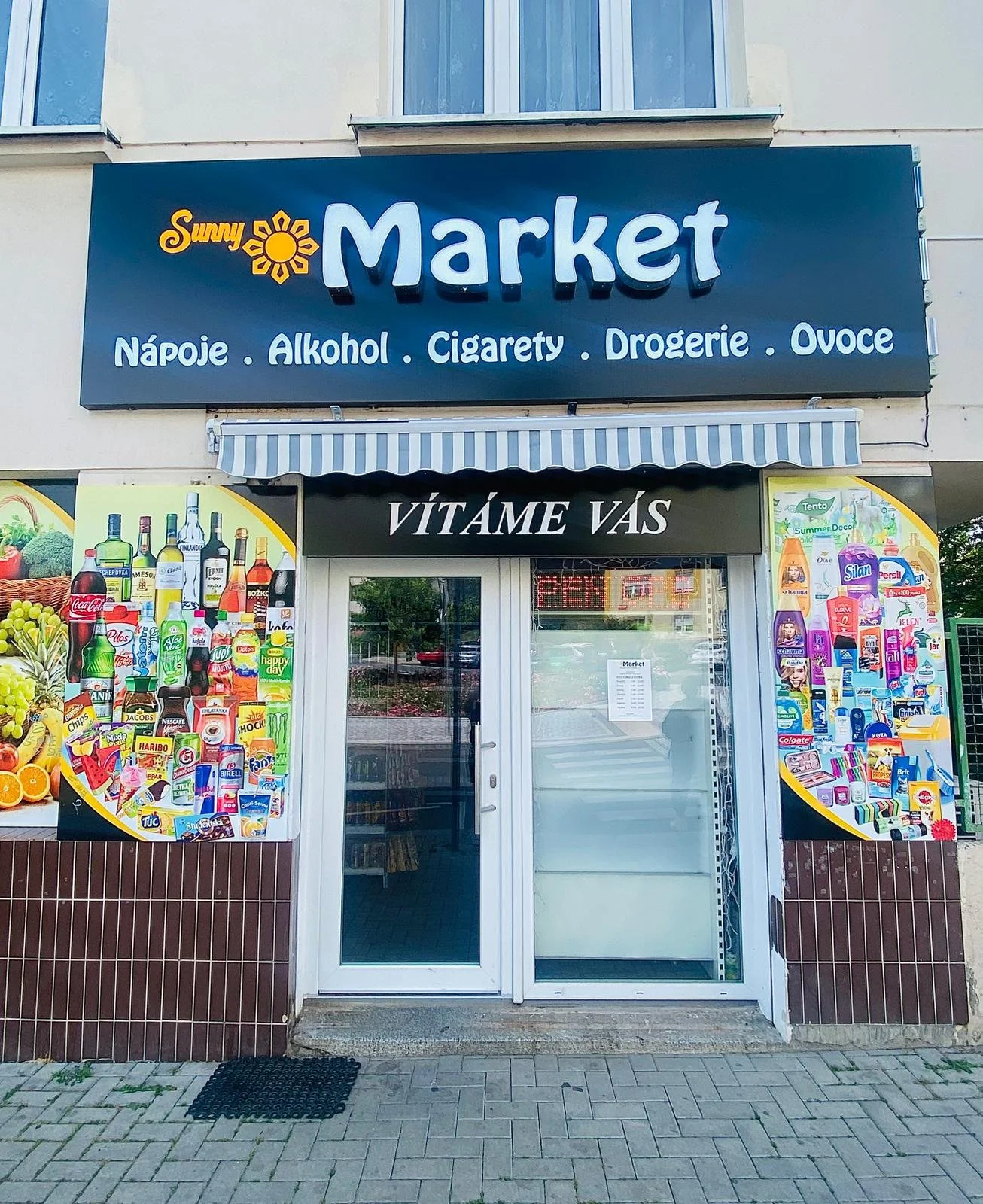 Sign for Sunny Market, a store selling beverages, alcohol, cigarettes, groceries, and fruits, with colorful advertisements of various products outside