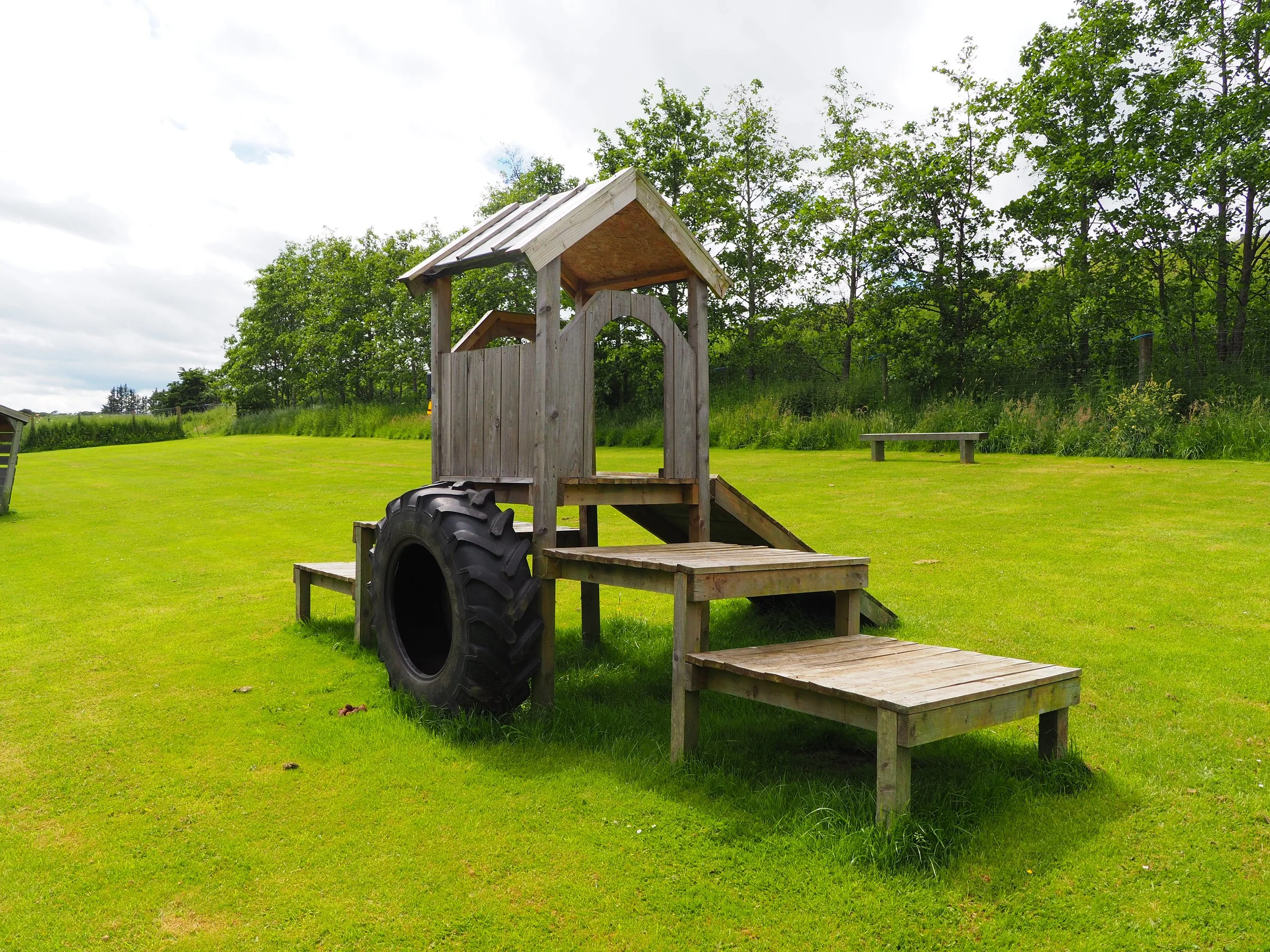 A wooden playground structure with steps leading up to a platform, a small roof, a slide, and a tire swing, set in a grassy yard with trees in the background.